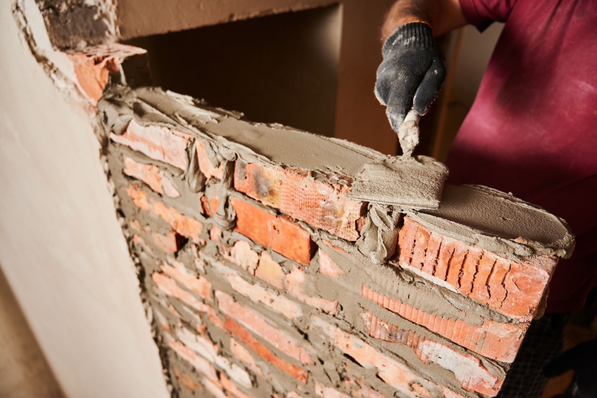 A person in a maroon shirt and black glove applies mortar to a row of red bricks while building a wall.