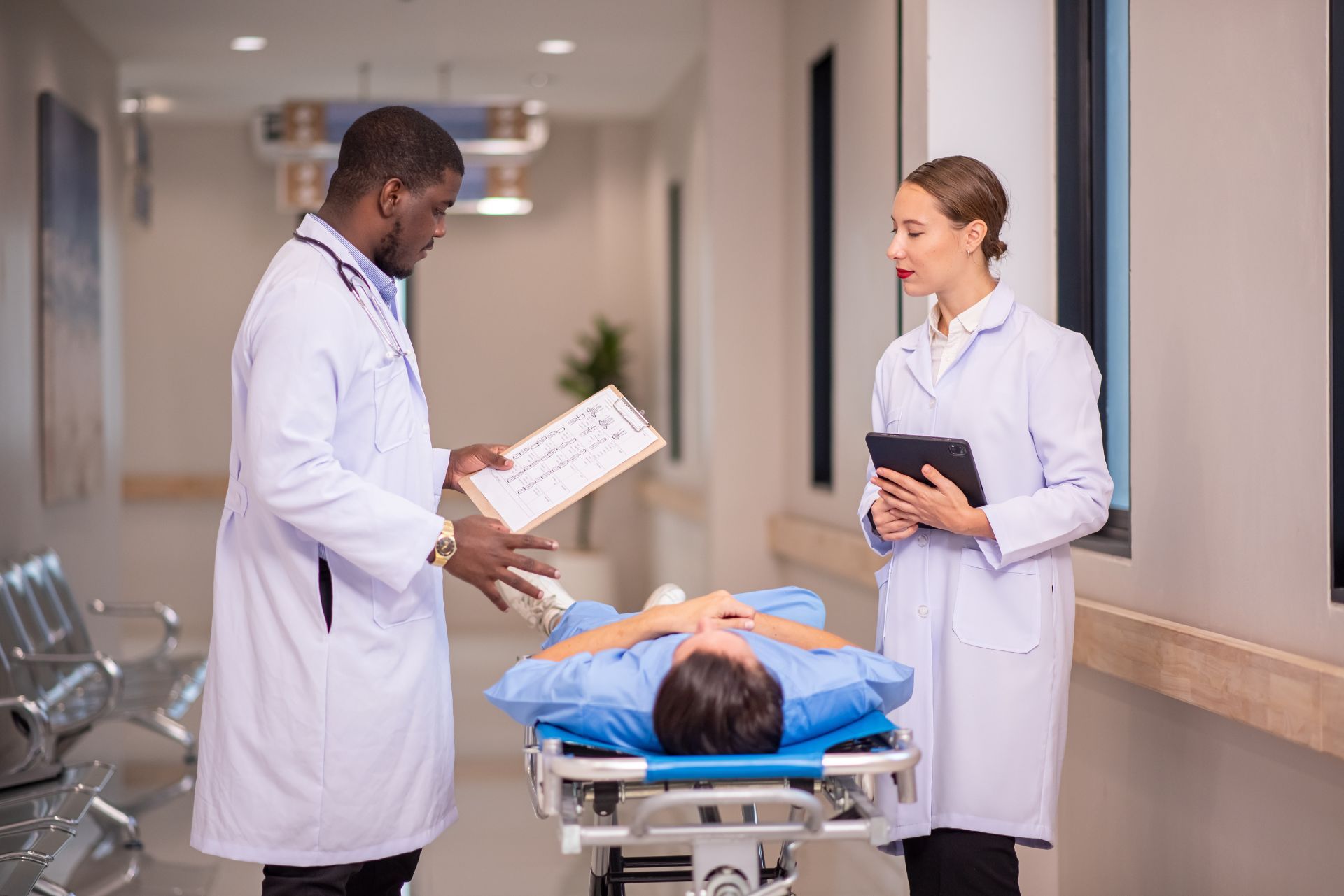 Doctors reviewing a patient on a hospital stretcher in a corridor, one holding a clipboard and the other a tablet