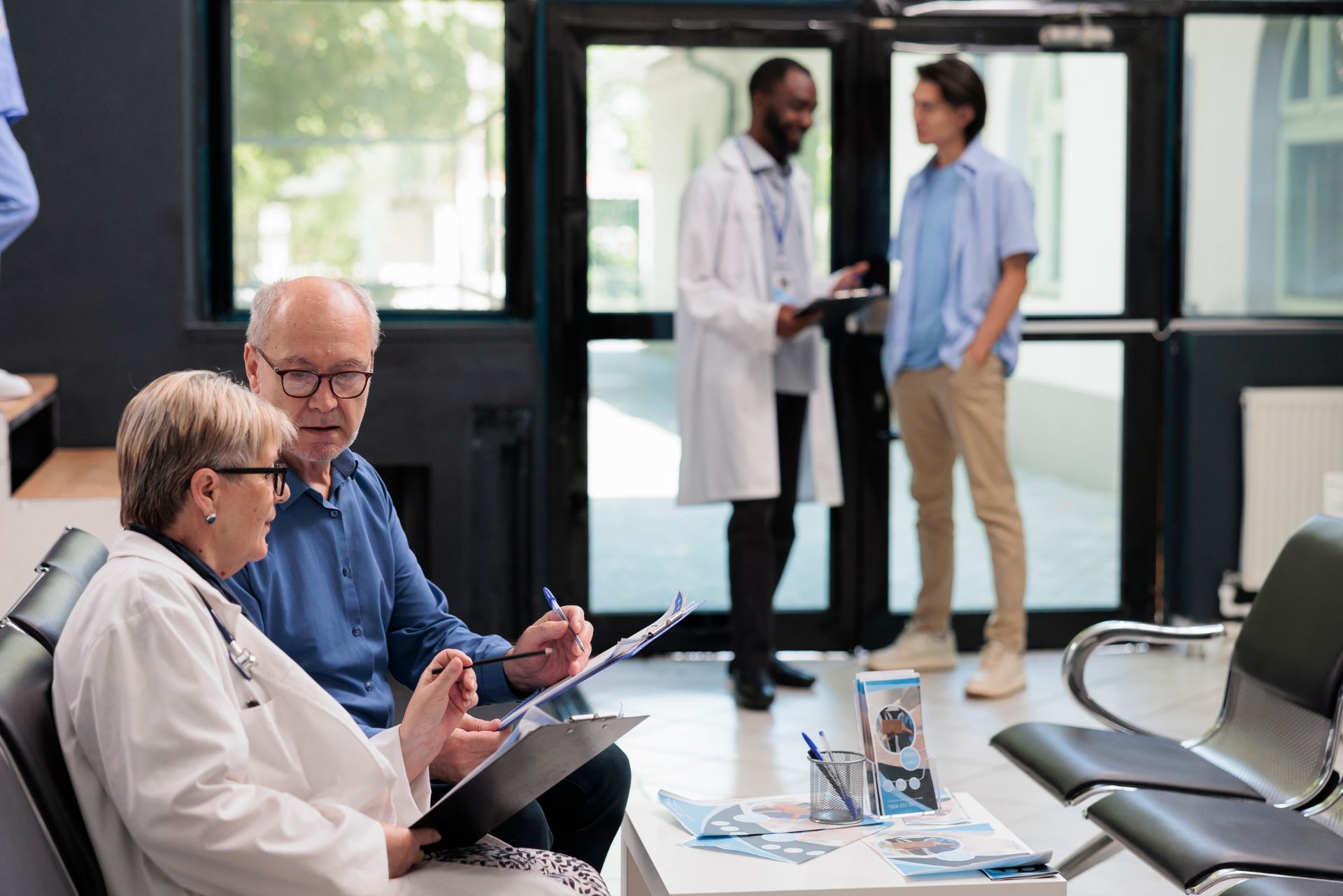 Patients and doctors in a clinic waiting area, with two people talking in the background.