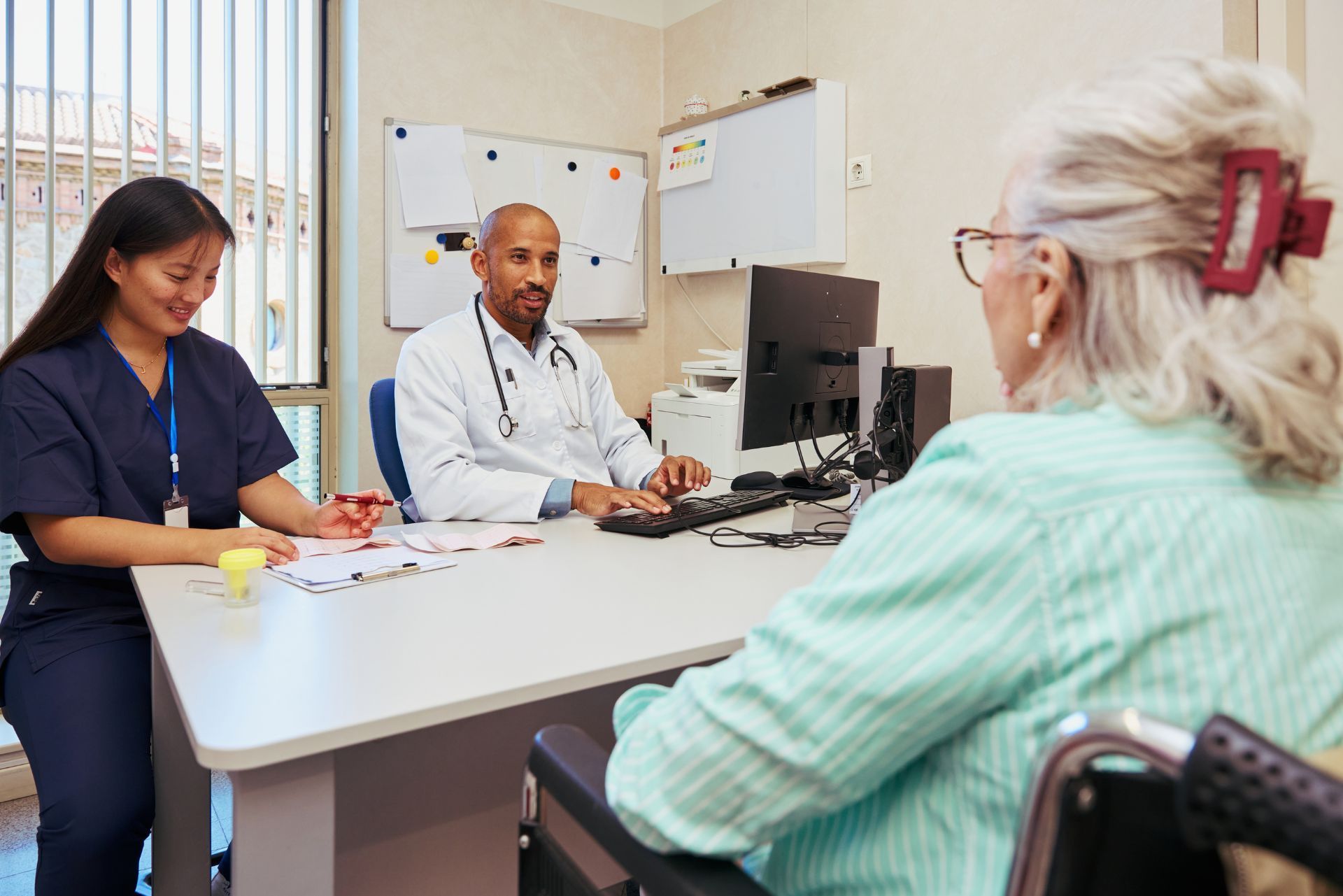 Medical consultation in a clinic, with a doctor and nurse speaking to a patient at a desk.