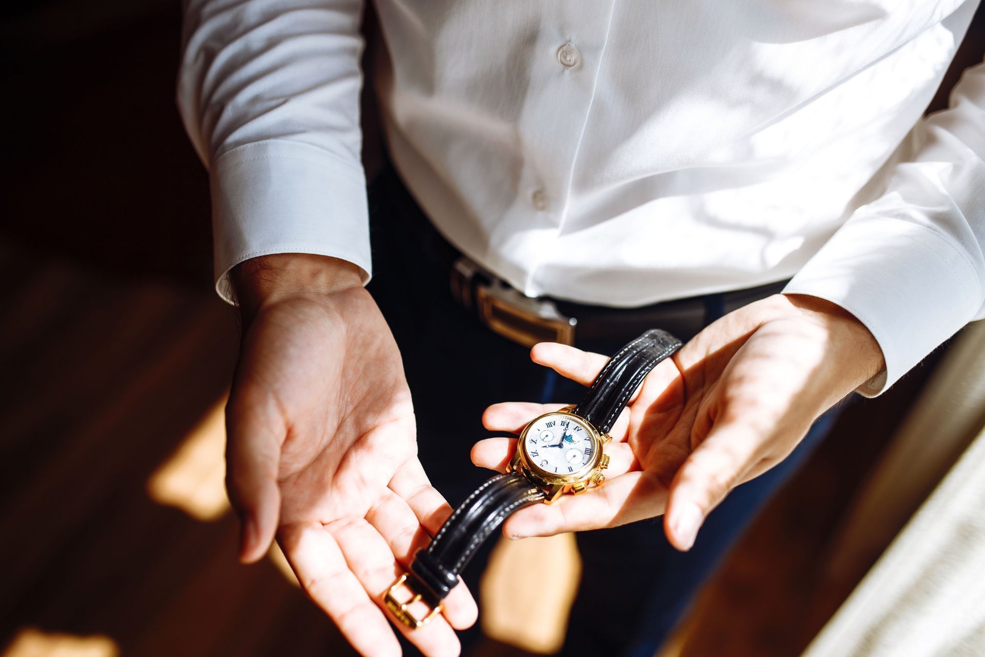 Hands holding a wristwatch with a black strap and gold case on a white shirt