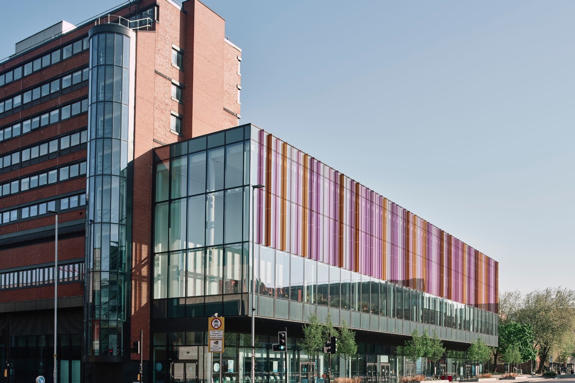 A tall brick building attached to a modern, multi-story glass structure with colorful vertical panels under a clear sky.