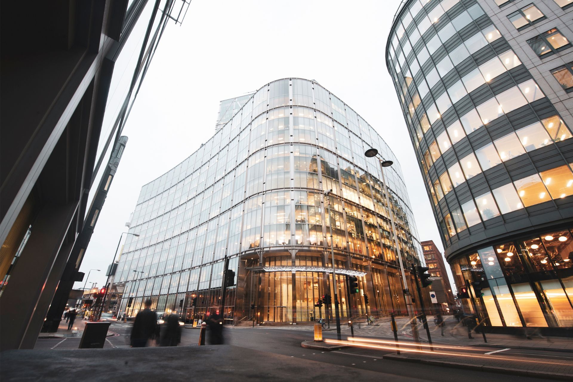 Curved glass office buildings stand on a city corner at twilight with glowing interior lights and urban street activity.
