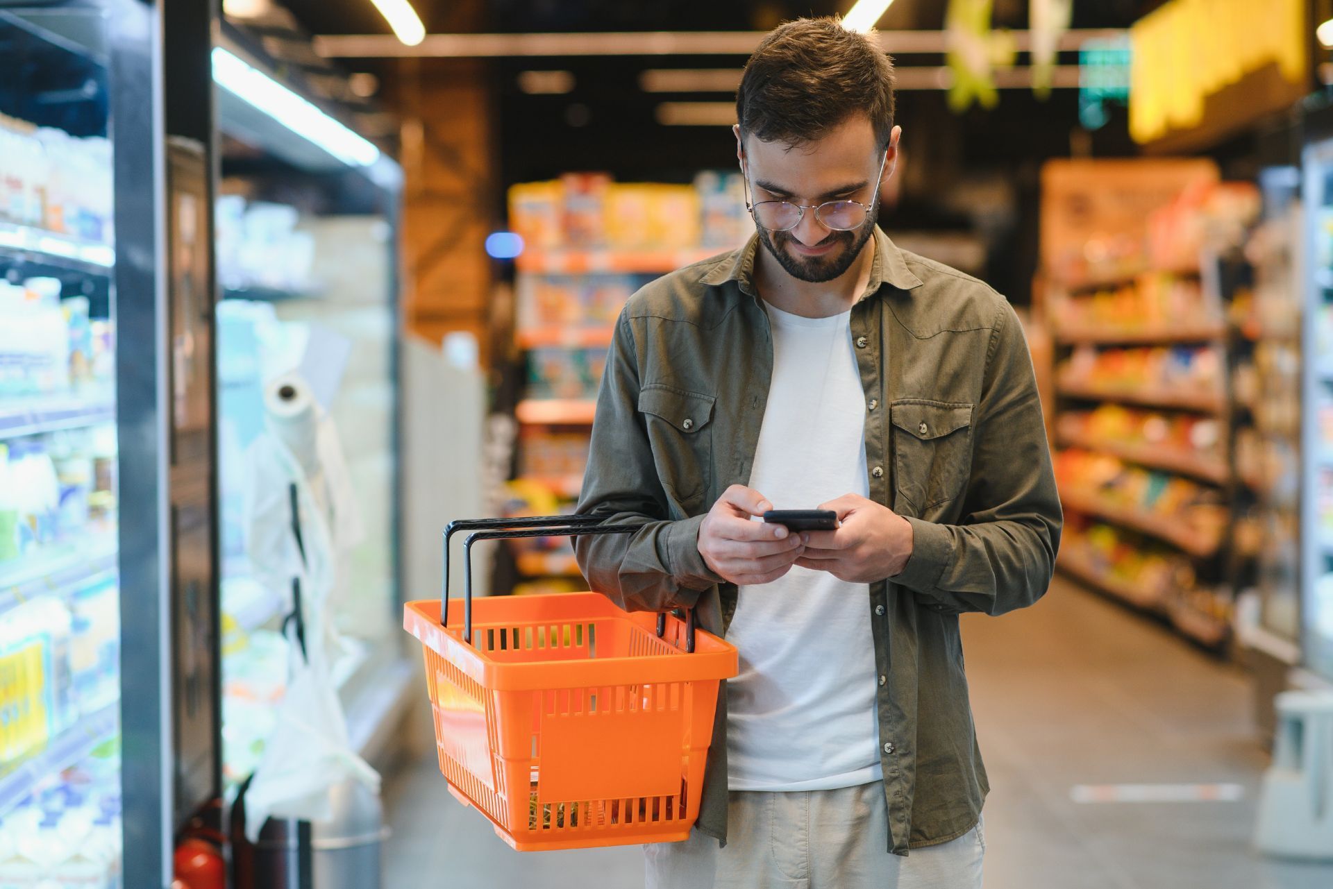 Man in a grocery store aisle looking at his phone while holding an orange shopping basket