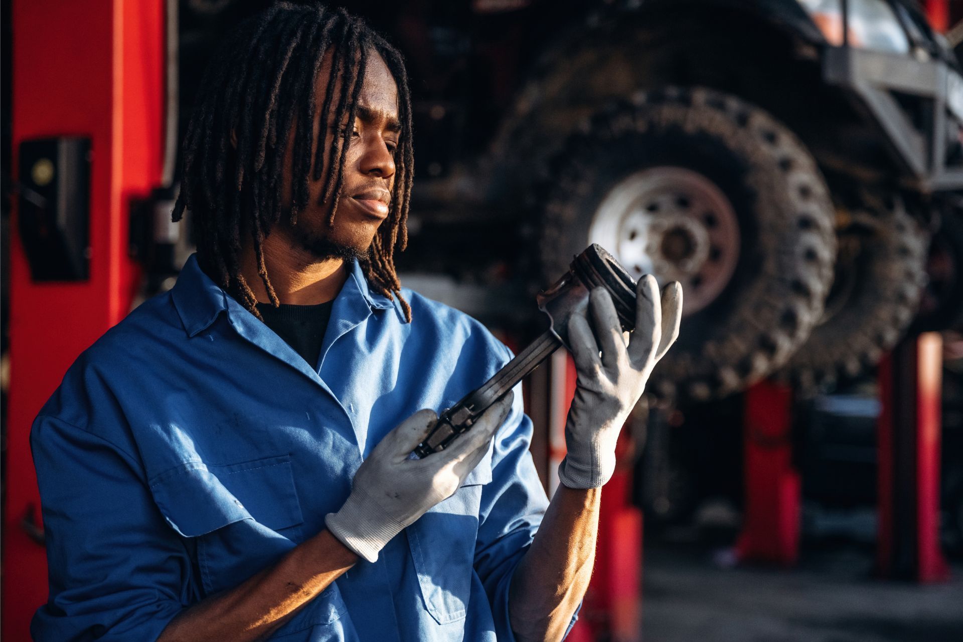 A mechanic in a blue uniform and work gloves inspects a metal engine component inside an auto repair shop.