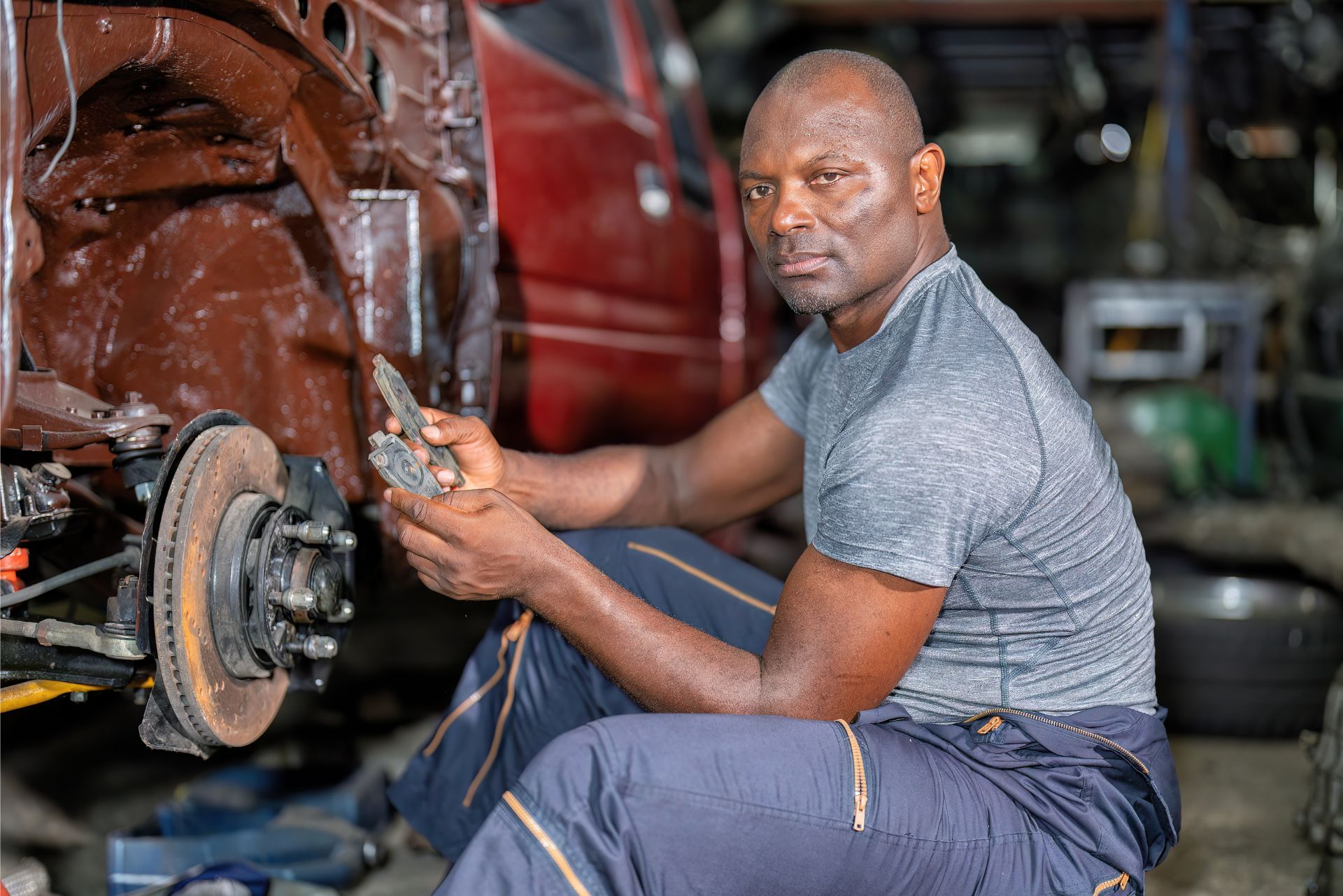 A mechanic in a grey t-shirt and blue trousers holds a brake part while crouching by a car in a workshop.