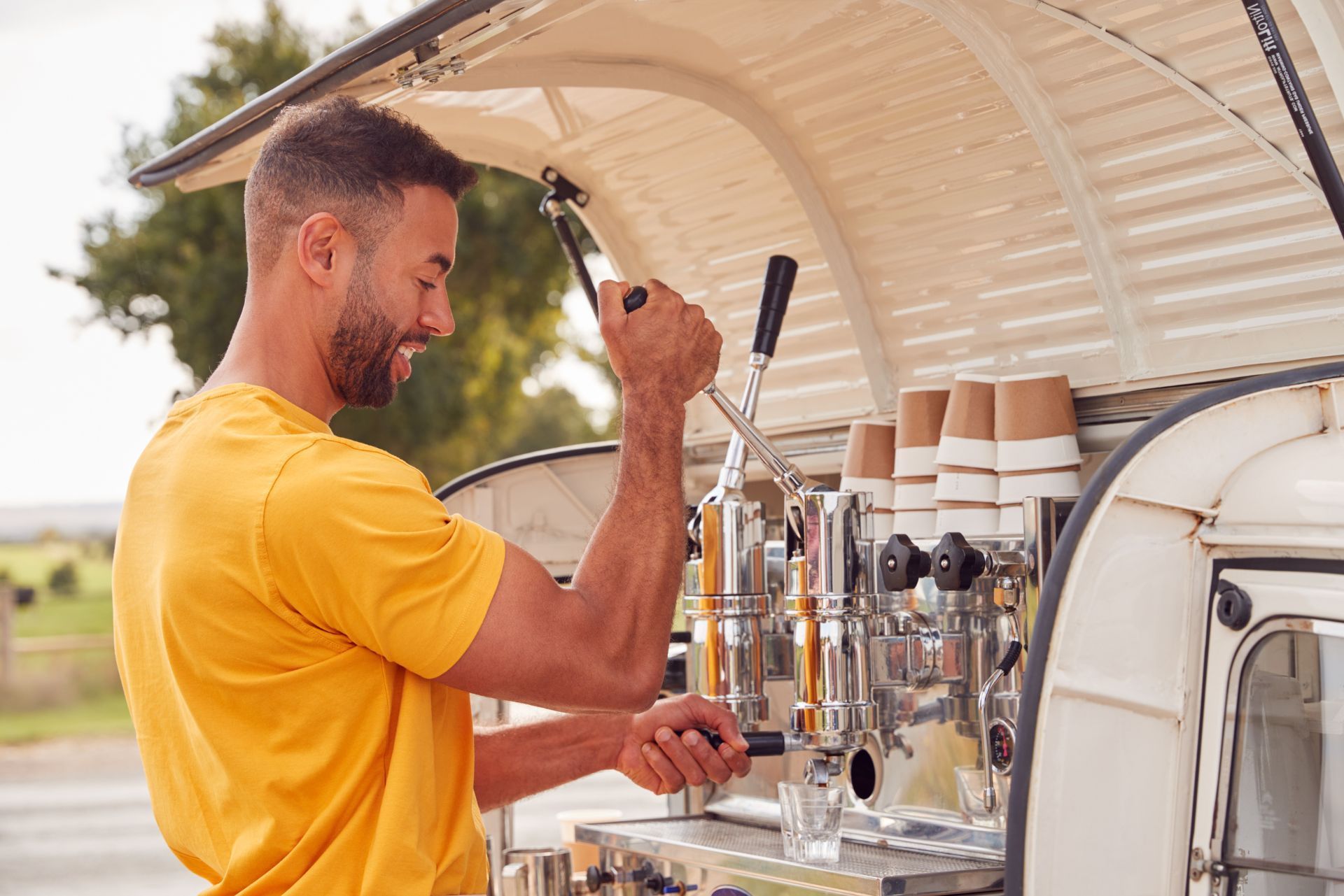 A smiling person in a yellow shirt operates a professional espresso machine inside a mobile coffee van.