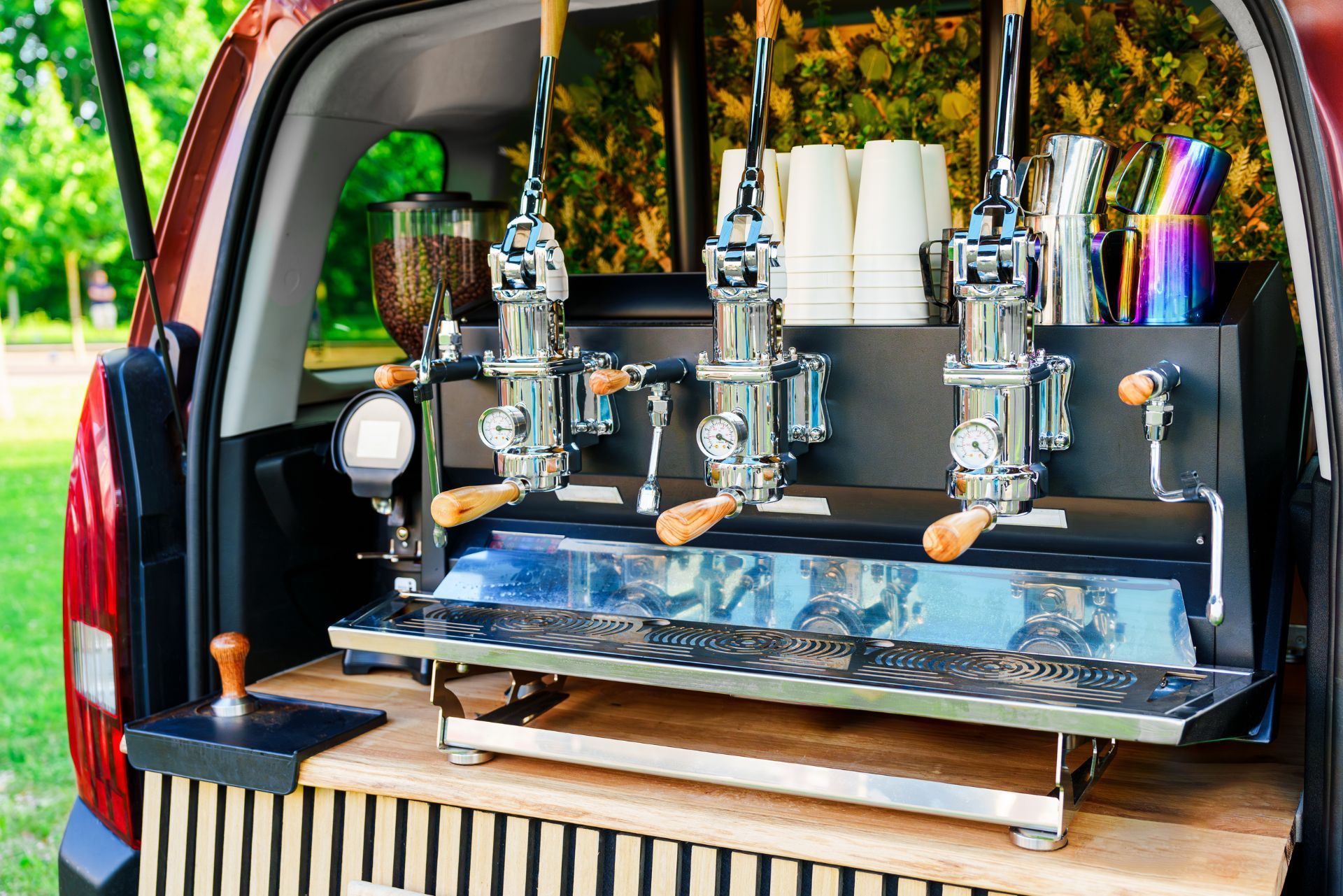 A three-group espresso machine installed in the back of a vehicle, featuring chrome levers, a wood counter, and cups.