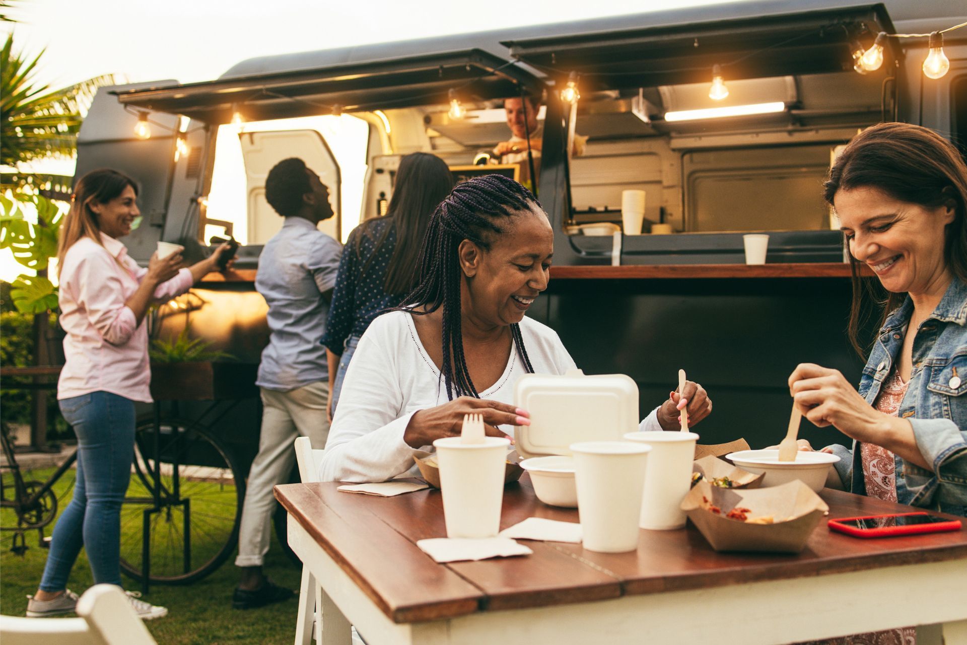 People eat at a wooden table in front of a food truck at sunset, with others ordering in the background.