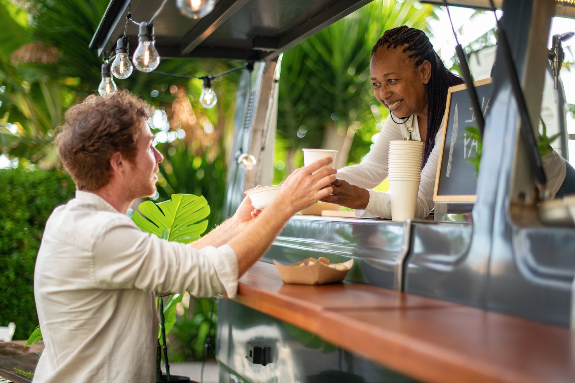 A person receives a cup of coffee from a smiling server at a tropical outdoor food truck.