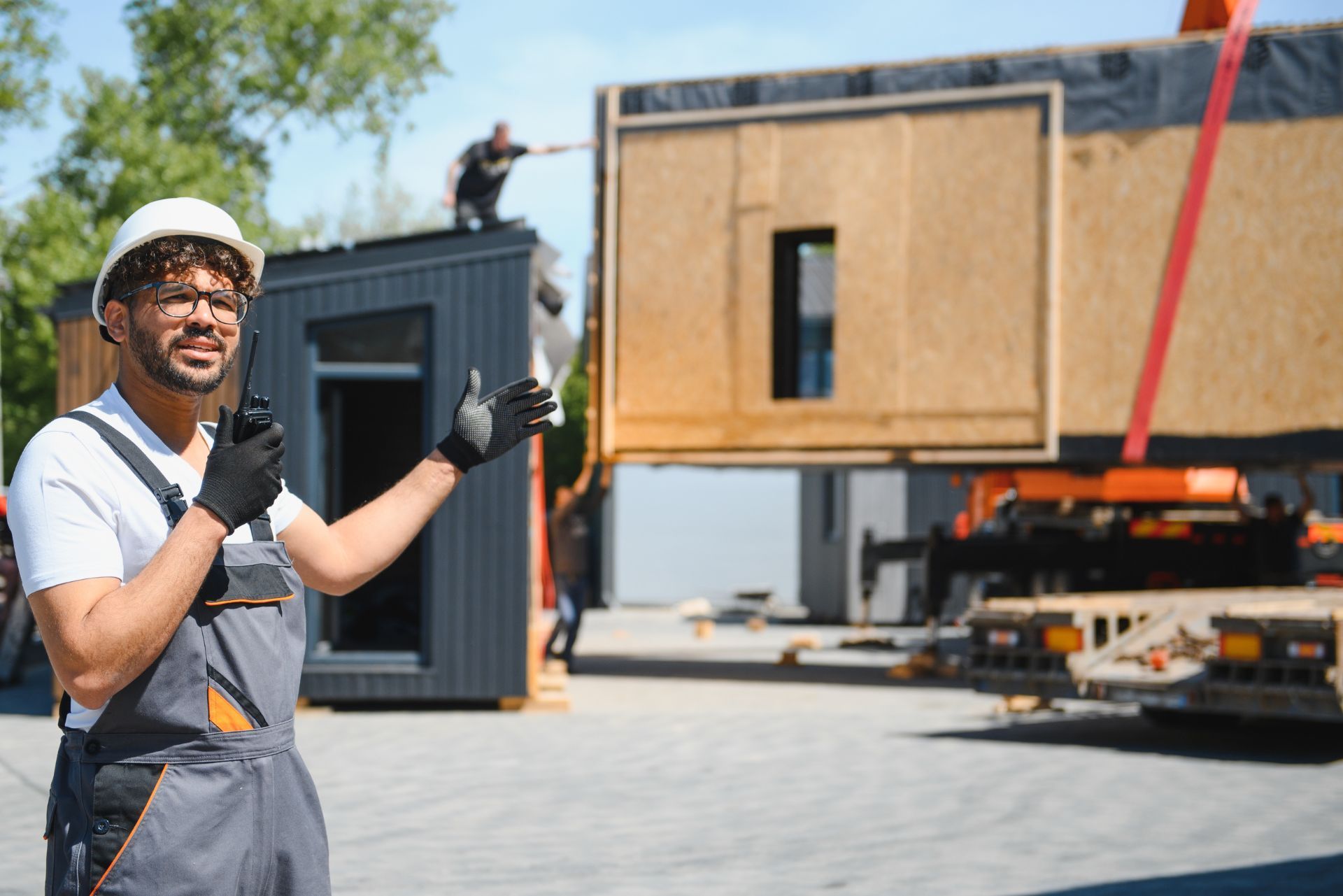 Worker in hard hat and gloves presenting a modular building being lifted by a crane in a construction yard