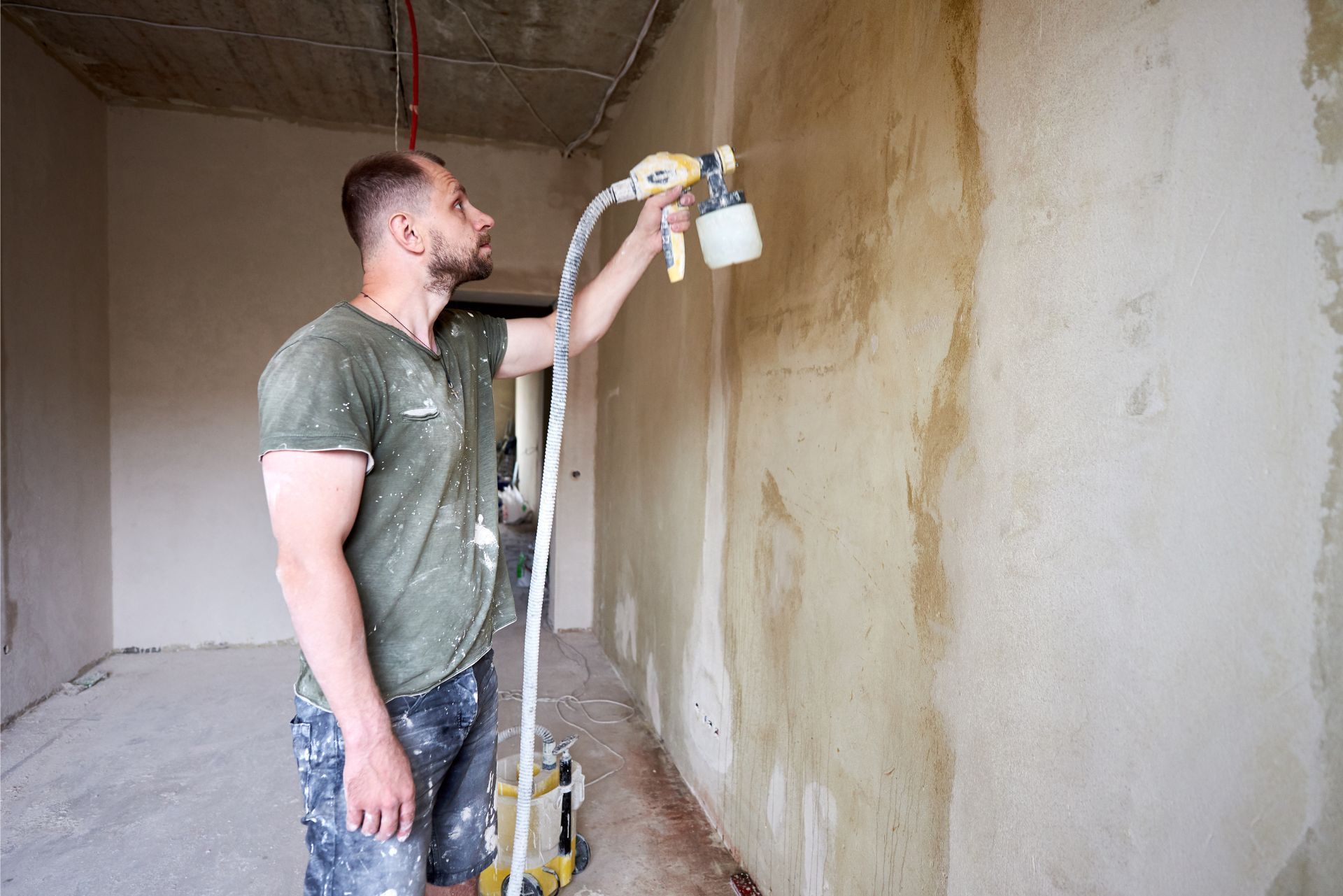 Worker spray-painting a wall indoors with a paint sprayer and extension pole