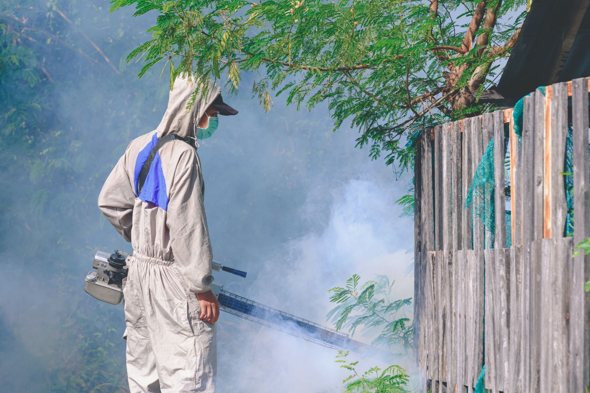 Person in white protective suit spraying smoke near a wooden fence and tree outdoors