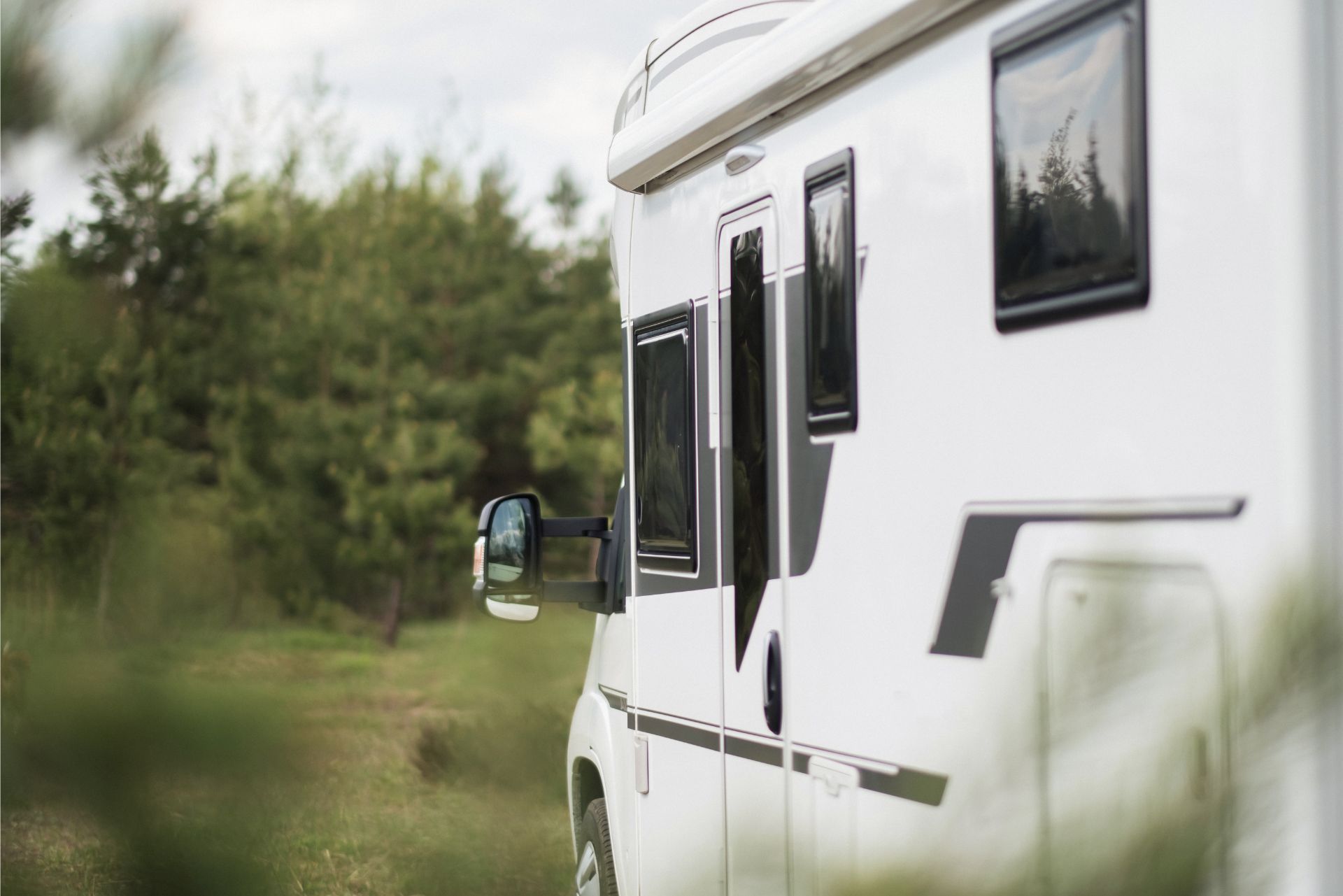 A white motorhome parked at the edge of a forest, viewed from the side.