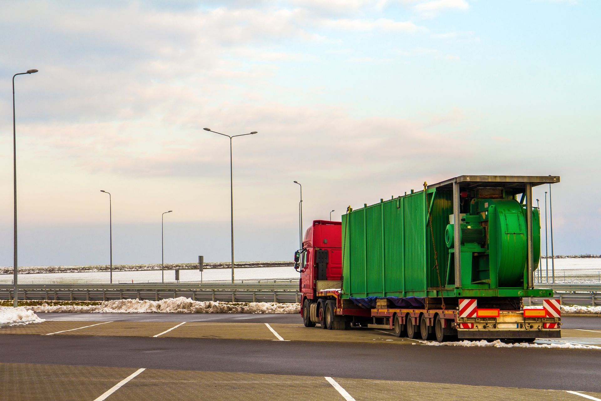 A red truck carrying a large, green industrial container parked in an open lot under a cloudy, winter-like sky.