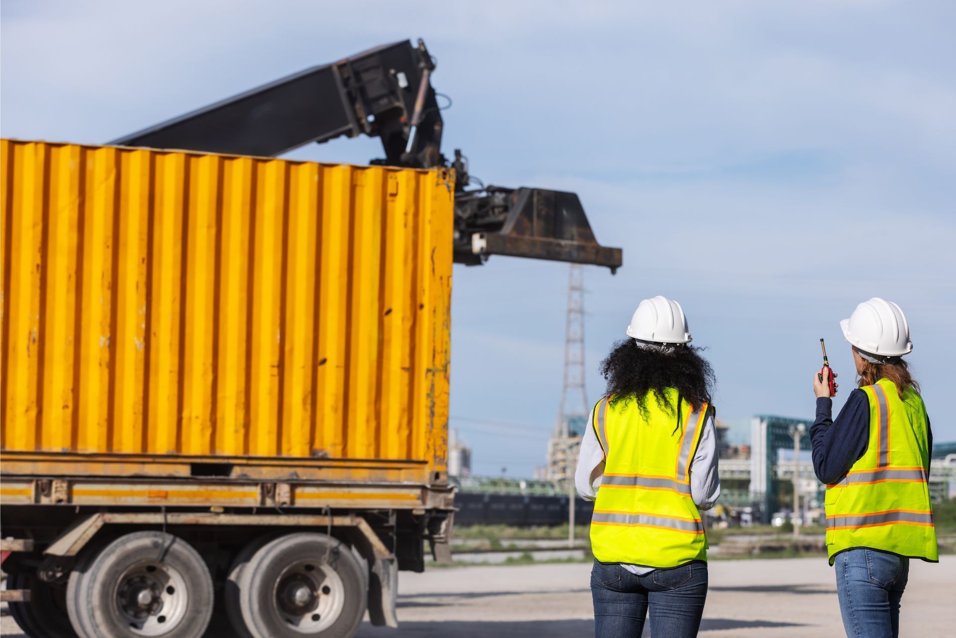 Two people in hard hats and high-visibility vests oversee a crane loading a yellow shipping container onto a truck.