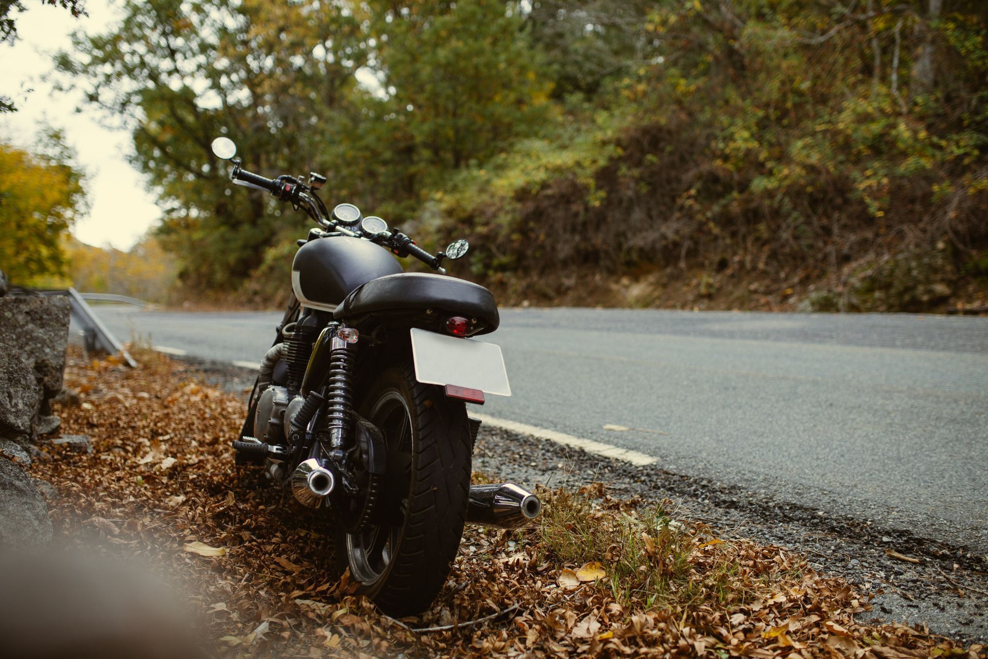 Black motorcycle parked on a leaf-covered roadside beside a curving asphalt road and wooded hillside