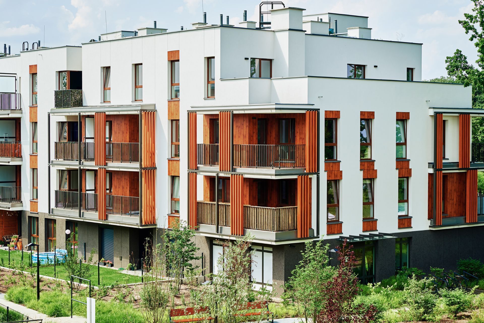 Modern white multi-story apartment building with wood-paneled balconies surrounded by green landscaping.