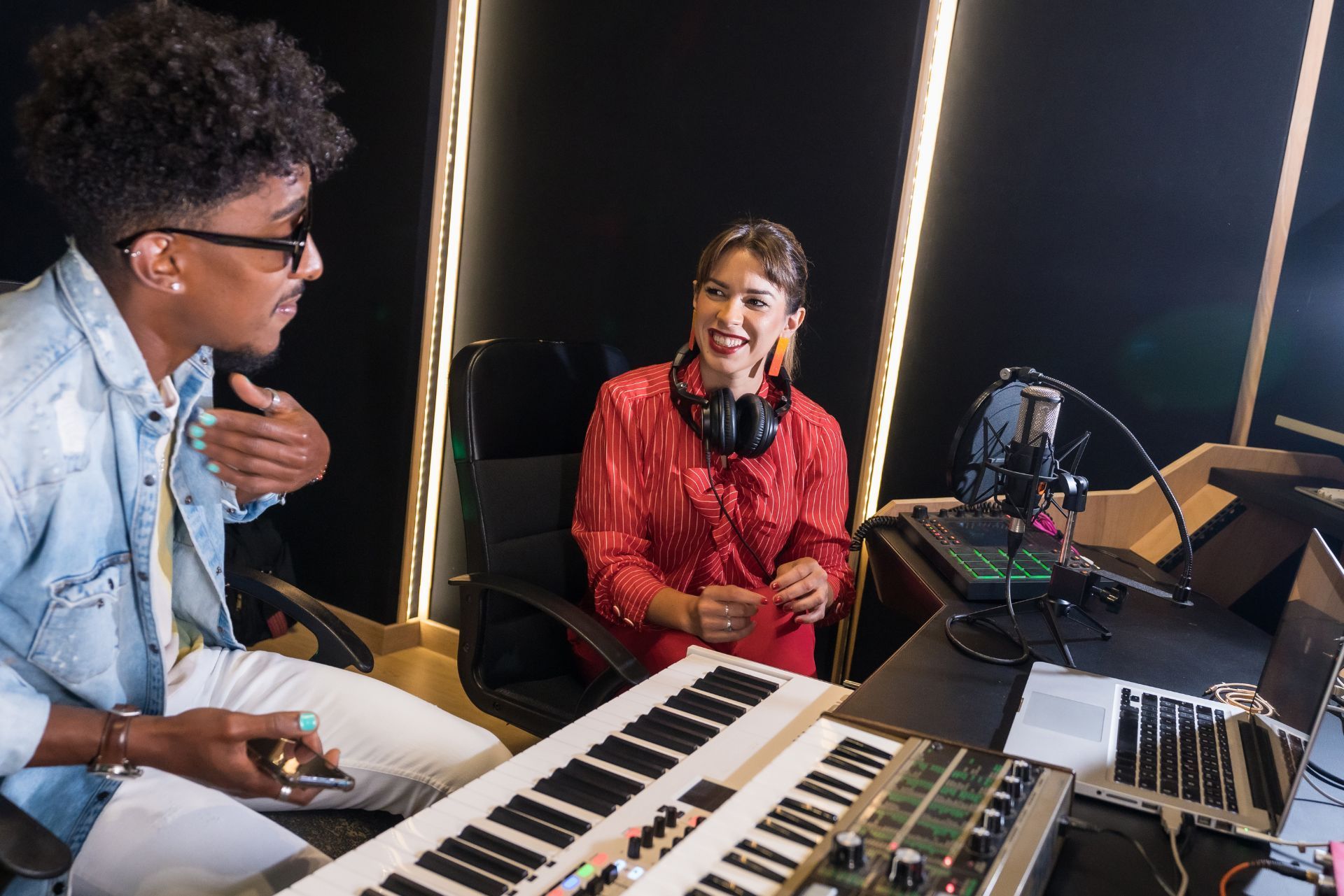 Two people in a music studio, smiling across keyboards and audio equipment