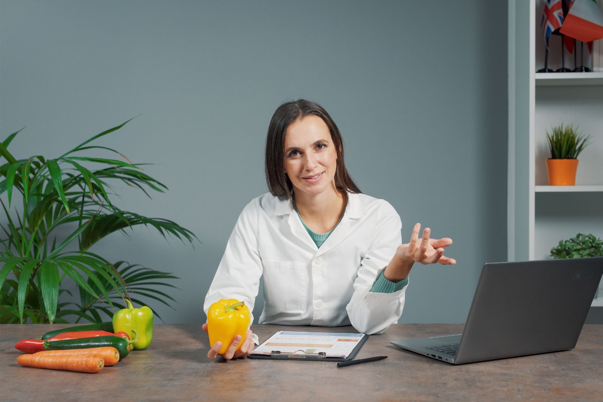 Woman in white coat at desk with laptop, vegetables, and a glass of orange juice, presenting with hand gesture