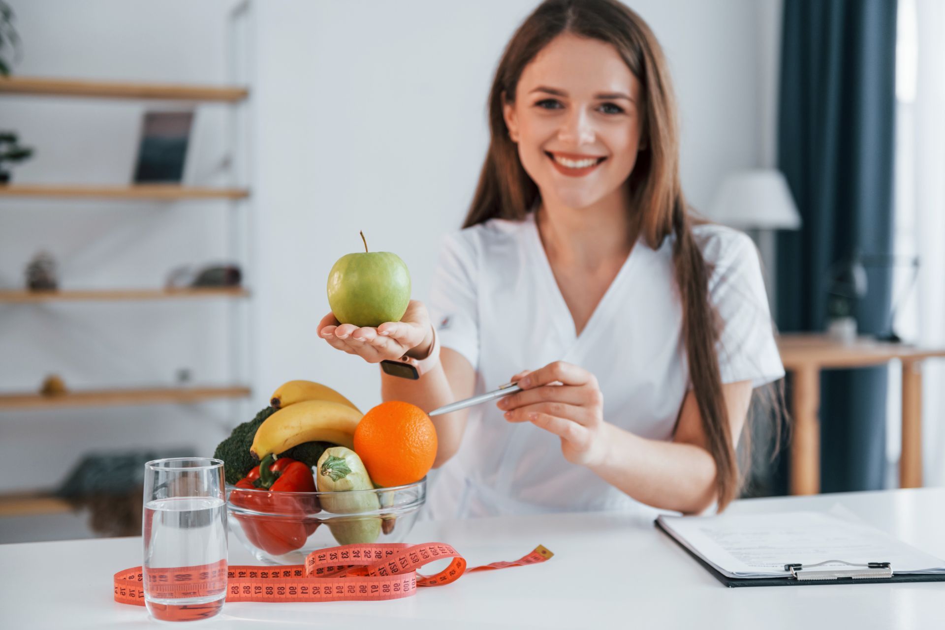 Woman presenting a healthy fruit bowl and green apple at a table with a laptop and measuring tape