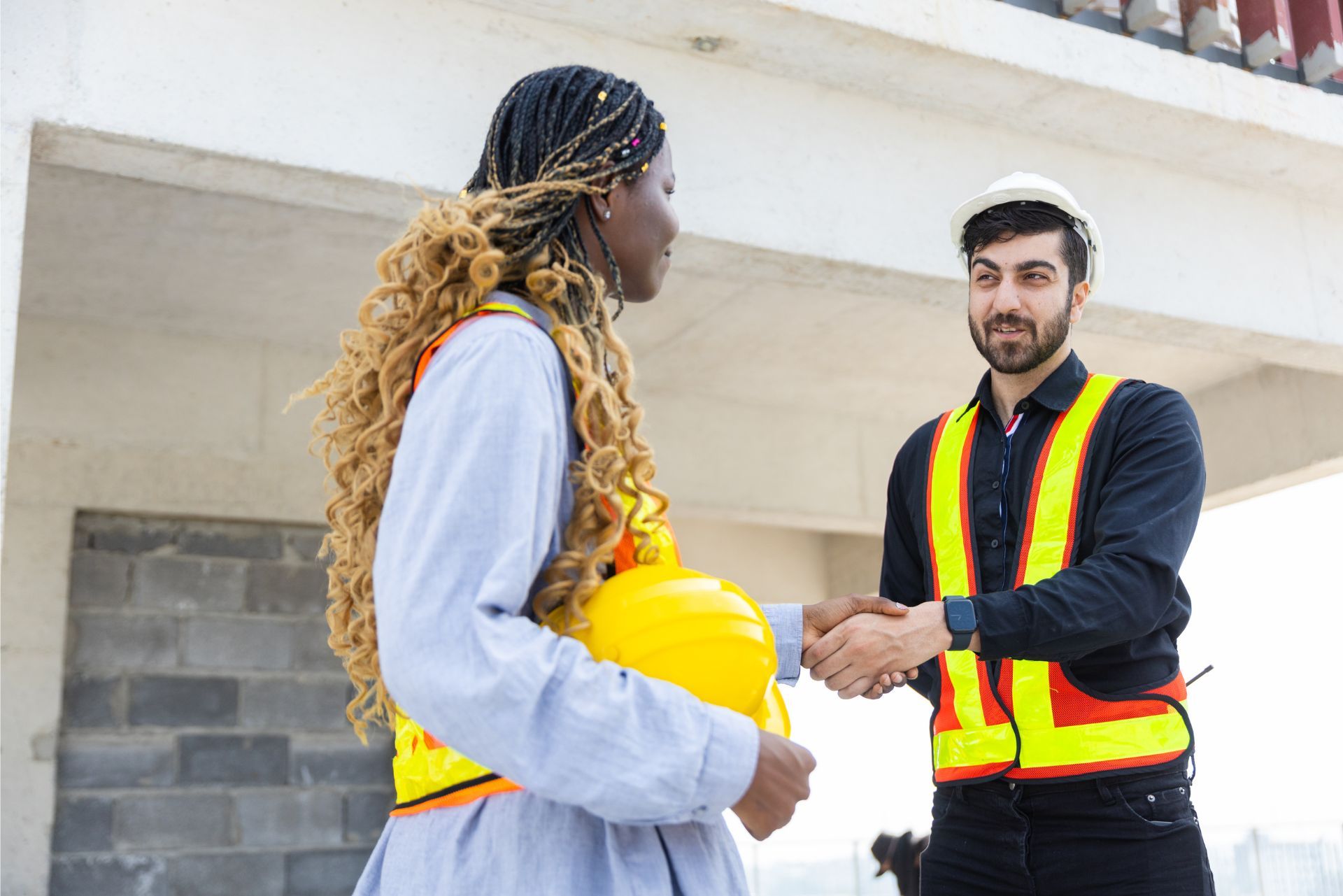 Two construction workers in safety vests talking at a building site, one holding a yellow hard hat