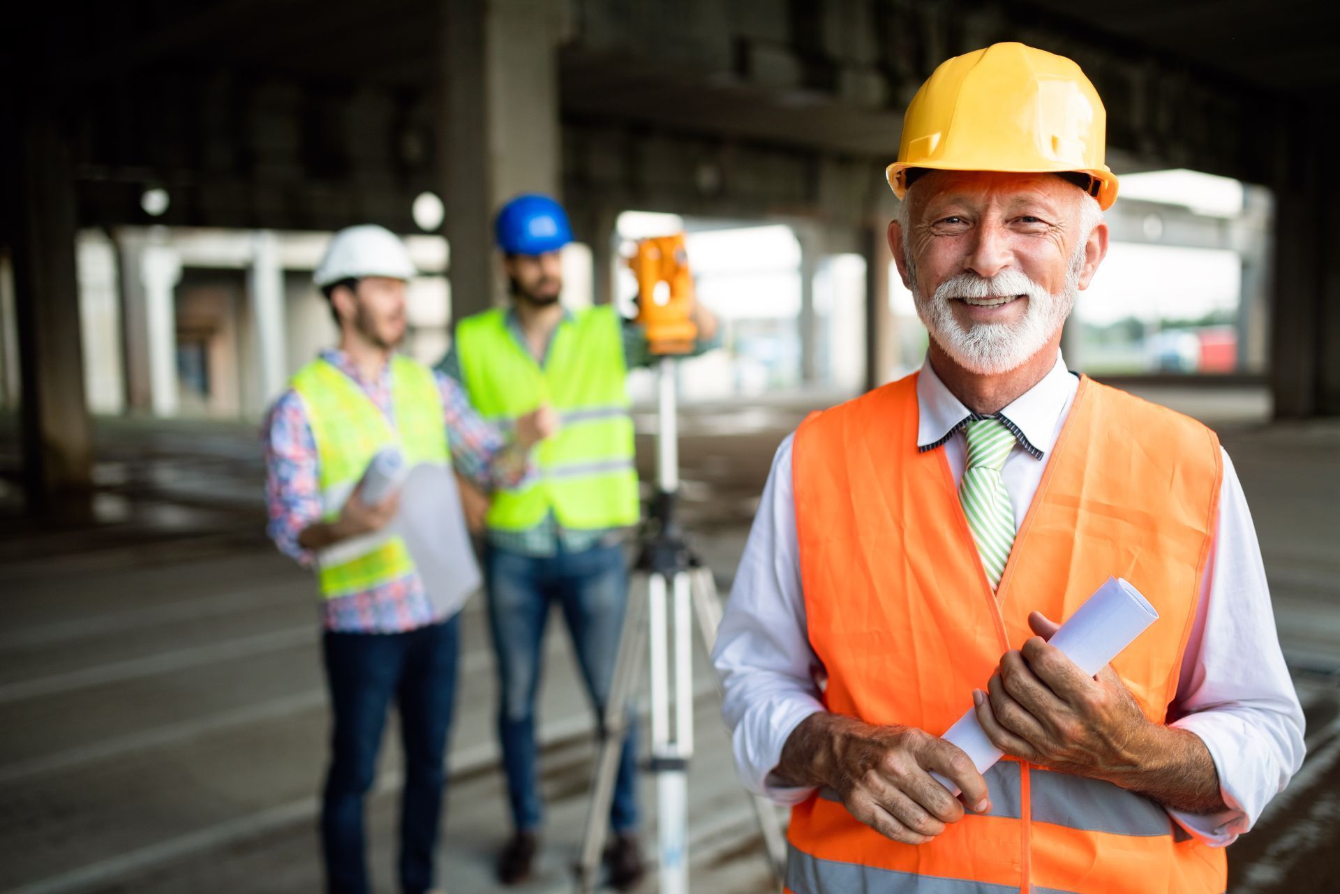 Construction team in safety vests and helmets at a worksite, with one man holding rolled plans.