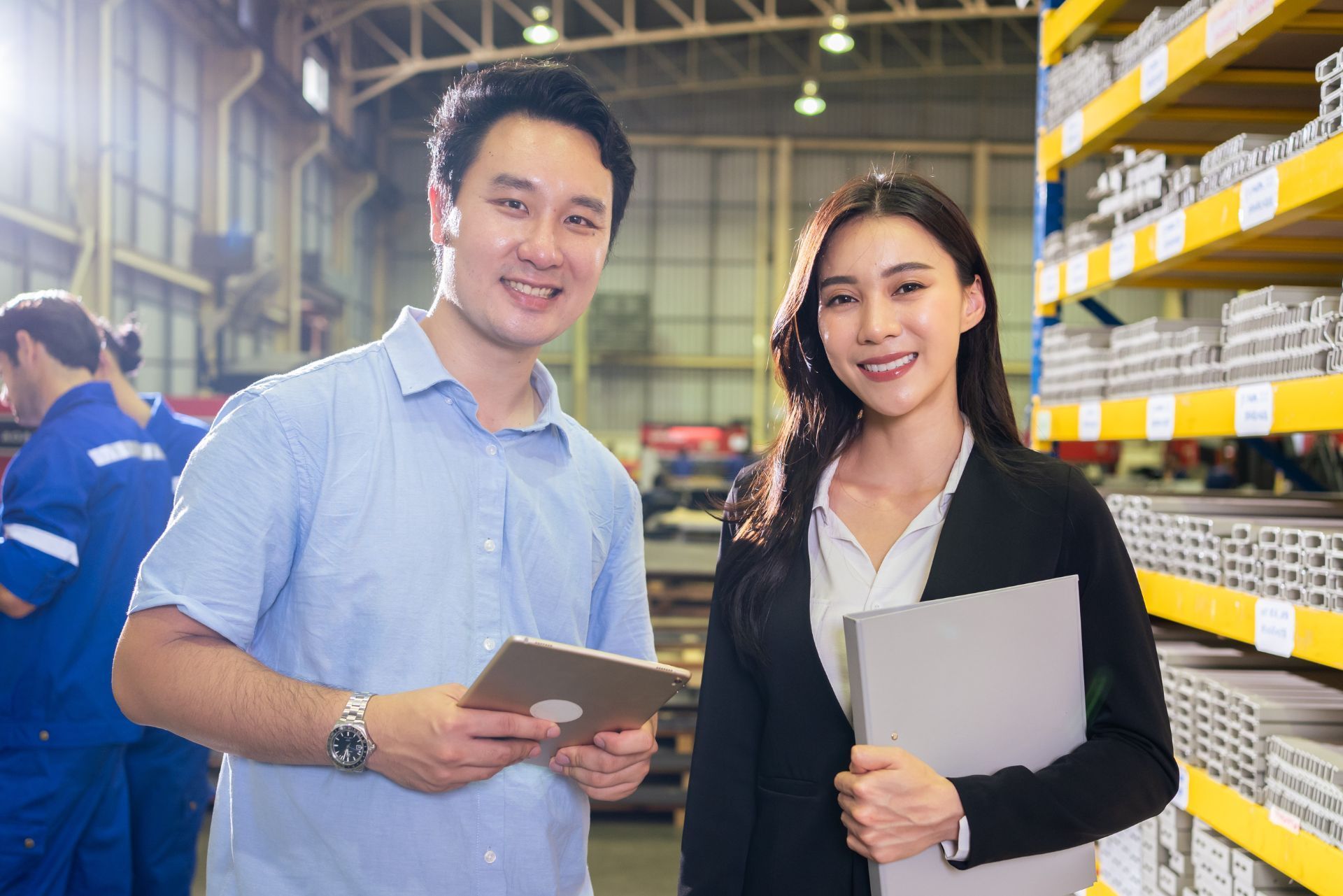 Two coworkers holding a tablet and folder in a warehouse aisle, smiling beside stocked shelves.