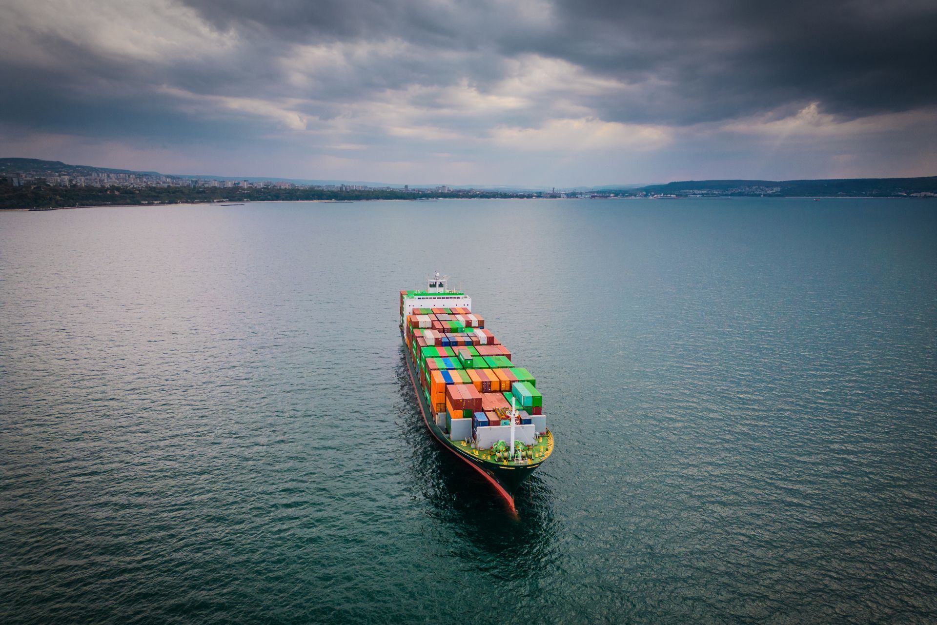 An aerial view of a container ship loaded with colorful cargo containers sailing on the open sea under a cloudy sky.