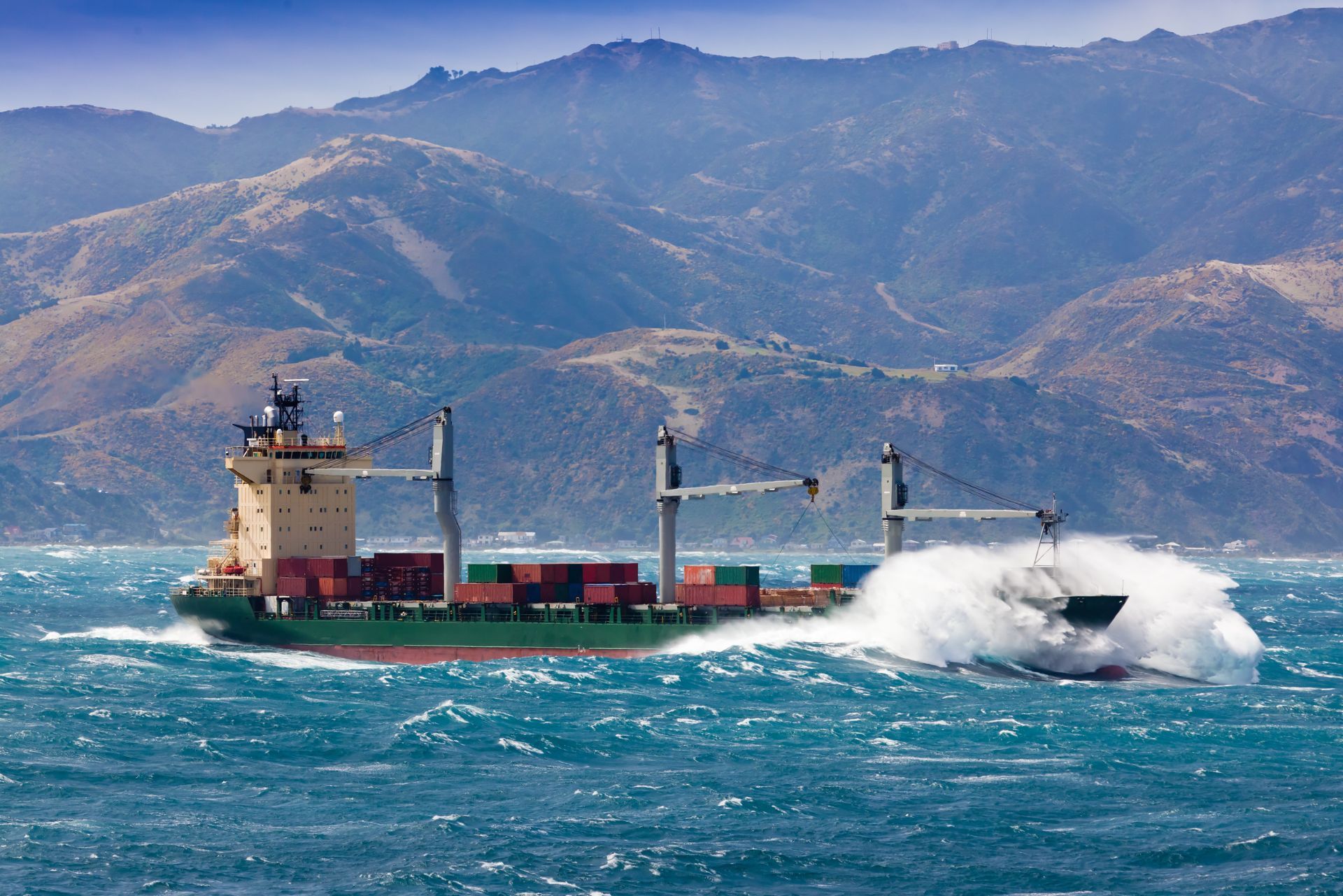 A green cargo ship traveling through rough, choppy water toward a mountainous coastline with white ocean spray.