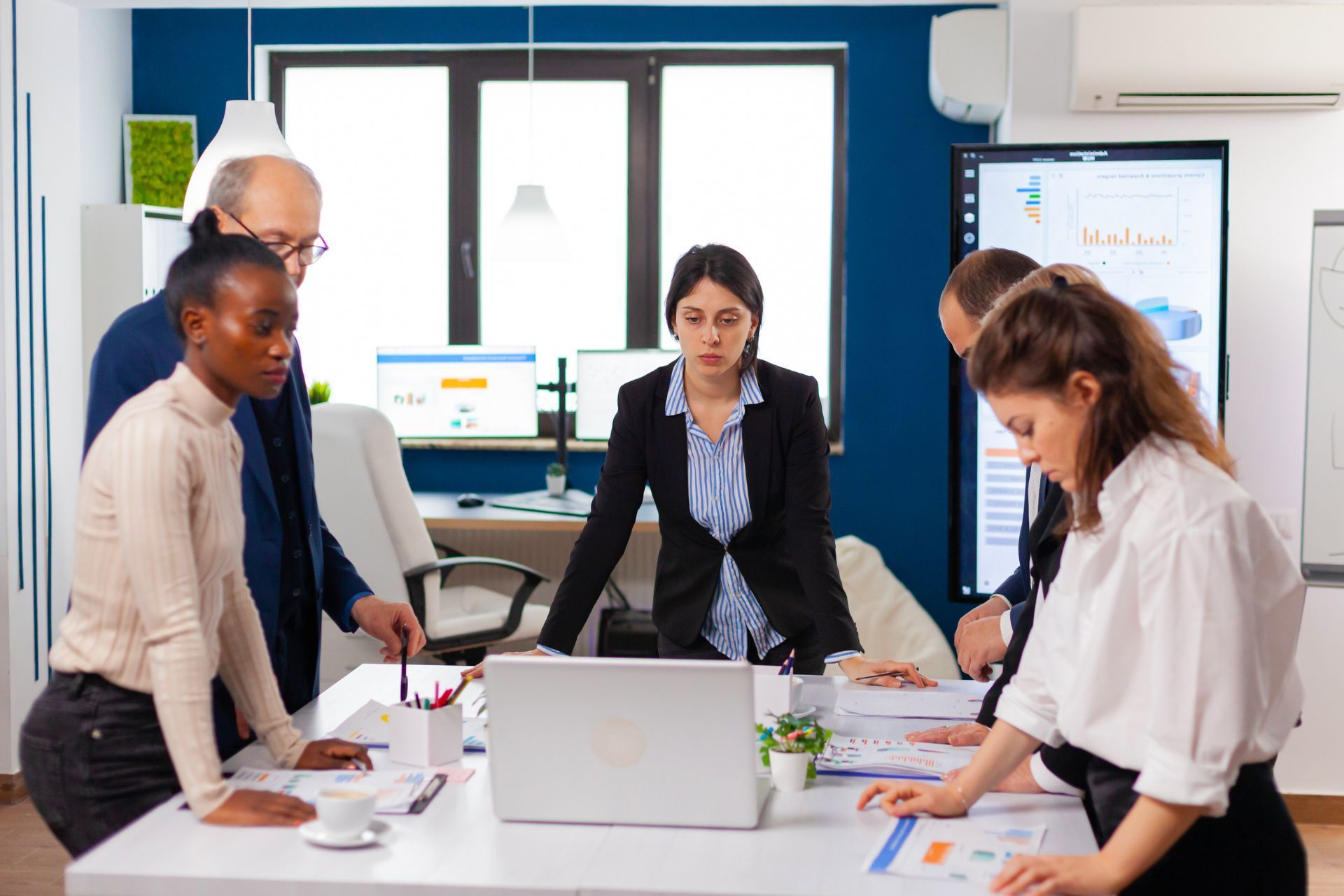 Colleagues in a meeting around a laptop in a bright office, reviewing documents and charts.