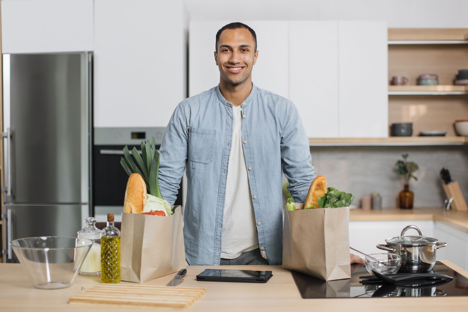 Man in a bright kitchen standing behind groceries, smiling beside a stove and countertop.
