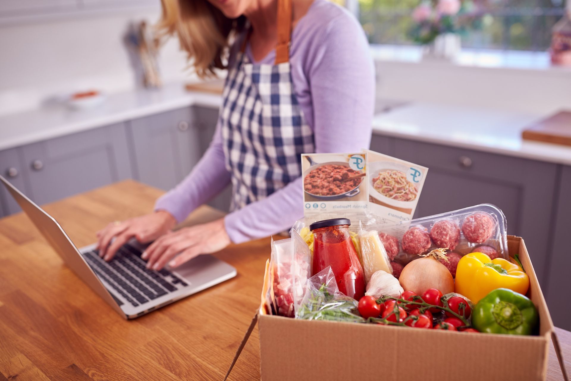 Person typing on a laptop beside a box of groceries in a bright kitchen