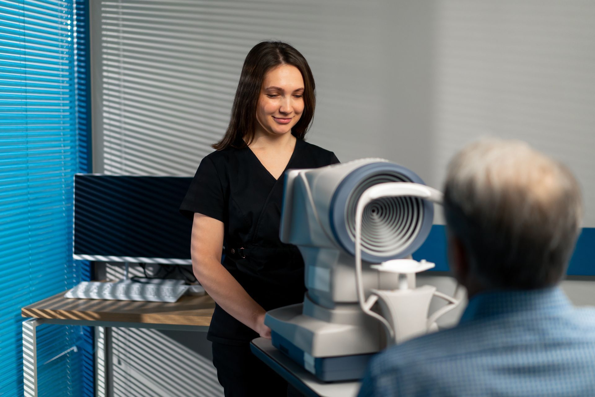Woman in black scrubs uses an eye exam device with a seated patient in a clinic.