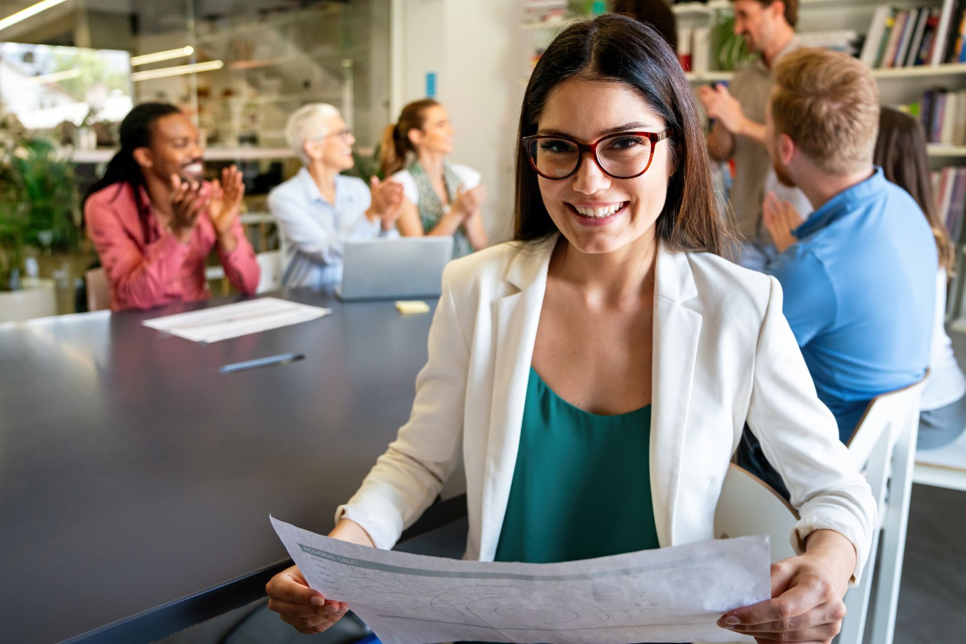 Smiling woman in glasses holding a paper in a conference room with coworkers seated at a table