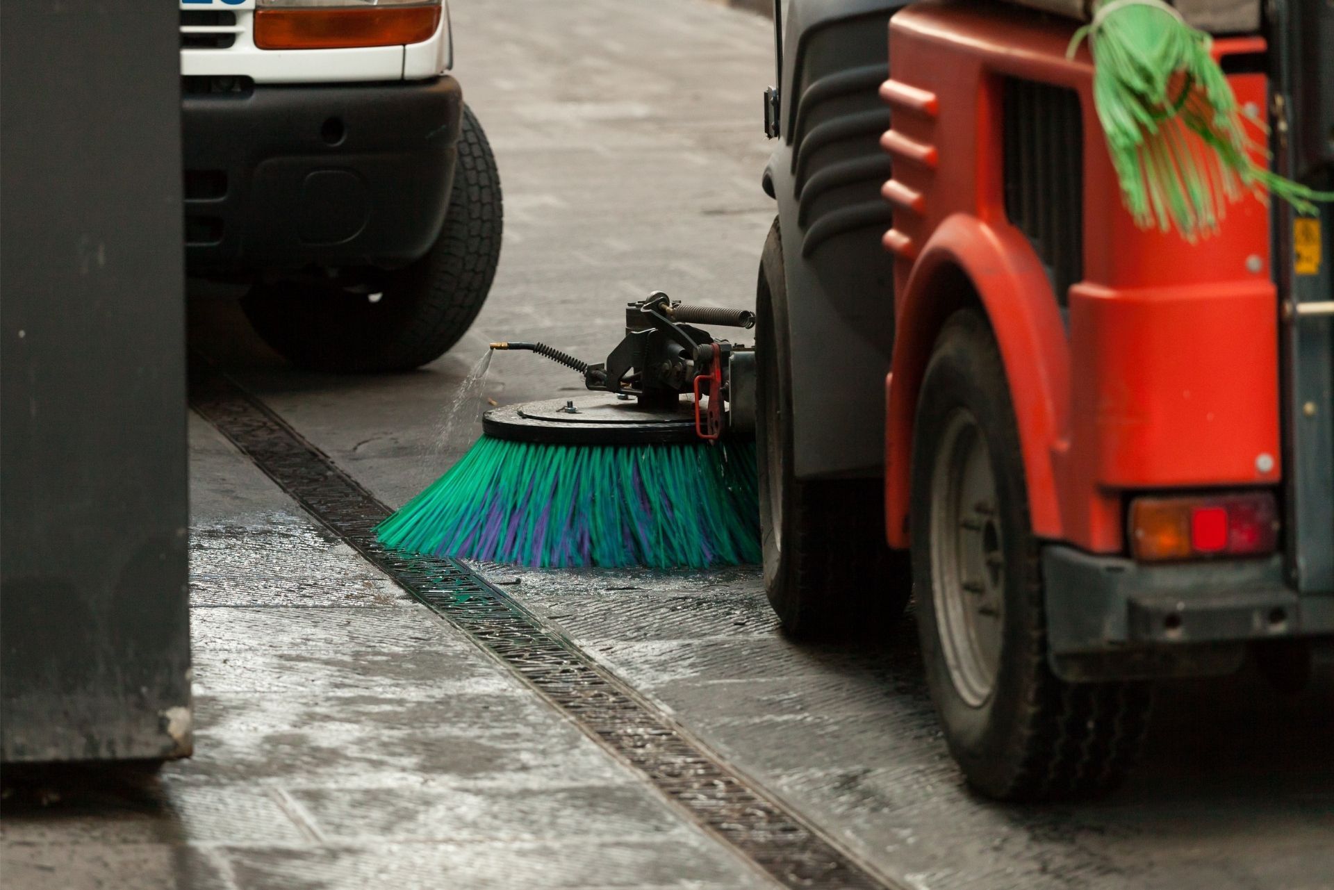 Street sweeper cleaning a wet curb beside parked vehicles and a red utility truck