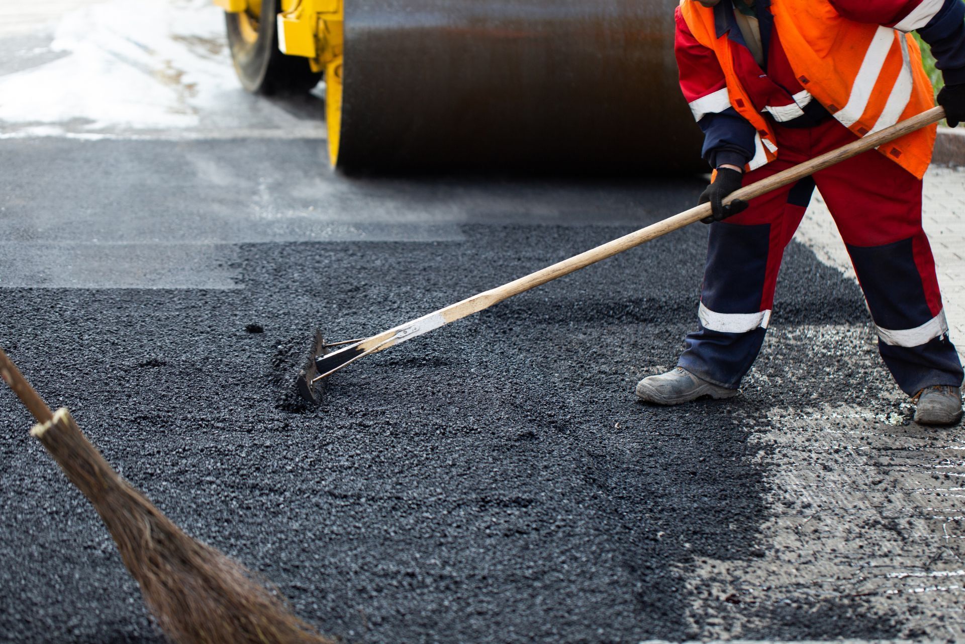 Worker spreading hot asphalt on road with a rake beside a paving roller
