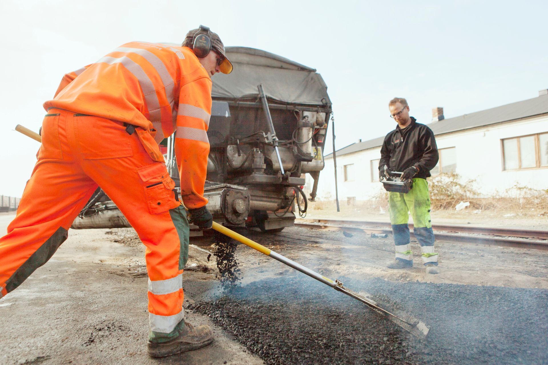 Road crew paving asphalt beside a truck, with one worker in orange gear and another standing nearby.