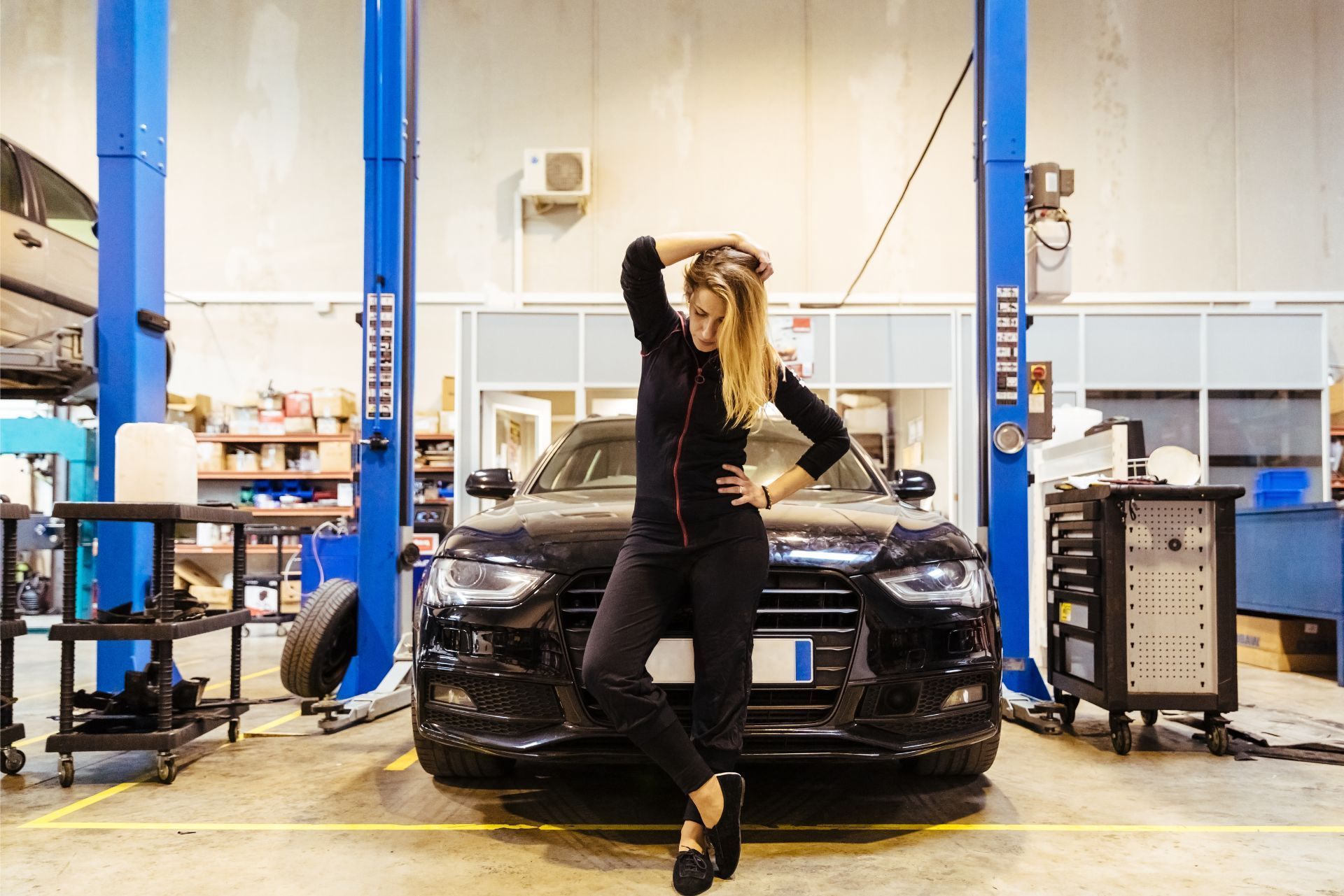 Person leaning on a black car in an auto repair garage with a blue lift.