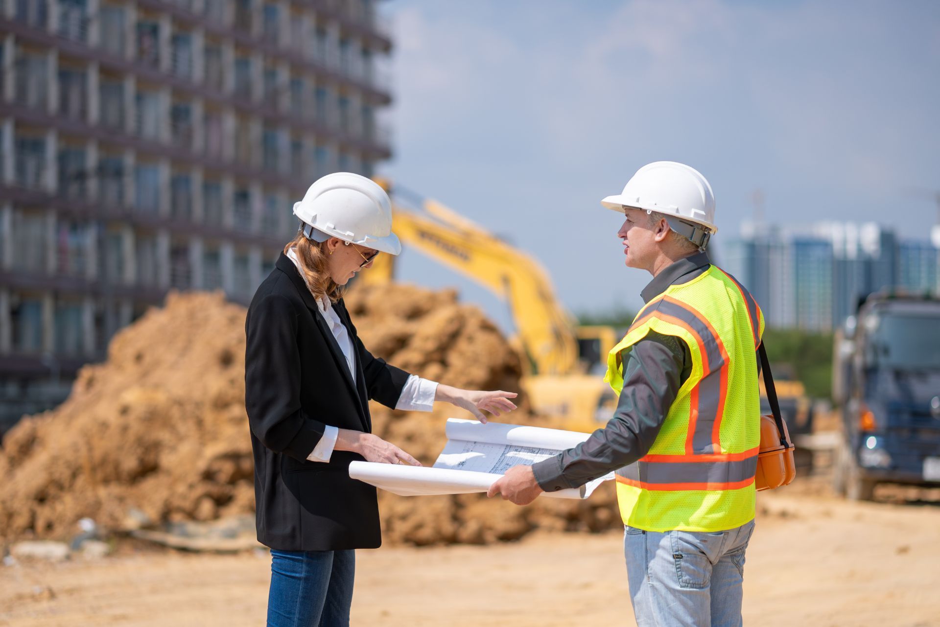 Two construction workers in hard hats reviewing blueprints at a building site with an excavator behind them
