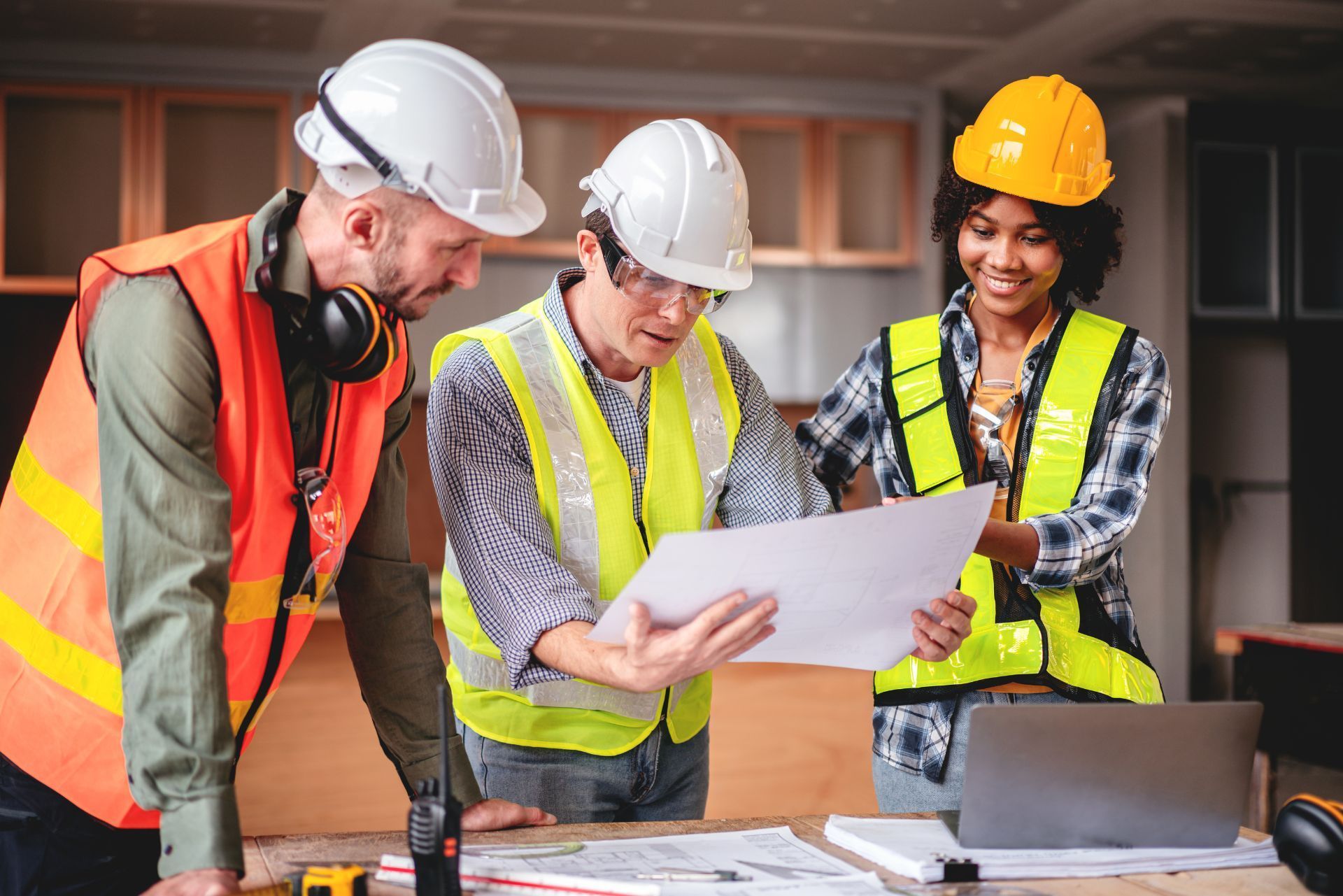 Three construction workers in hard hats review blueprints at a job site.