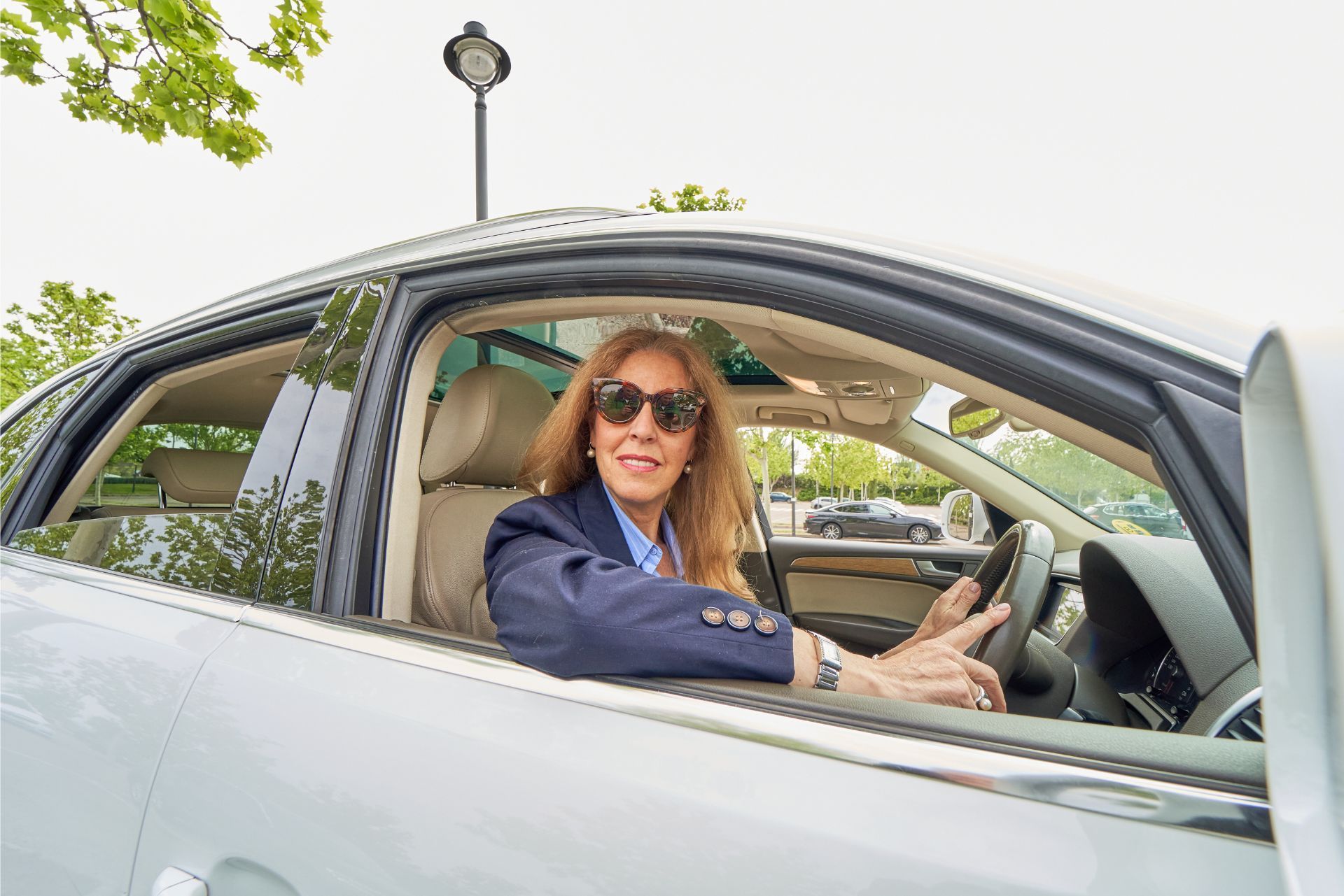A person wearing sunglasses and a navy blazer drives a car on a bright day.