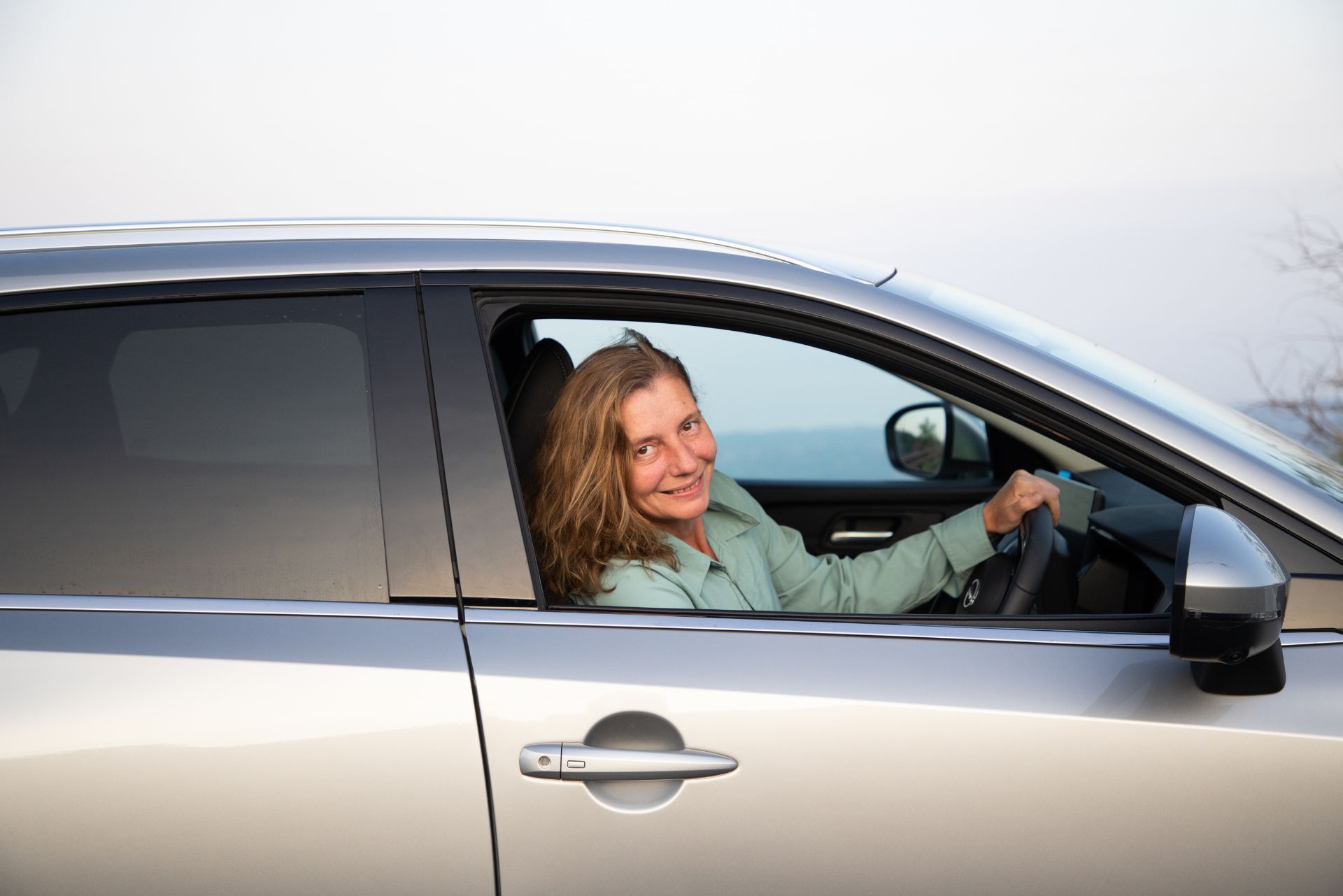 A person smiles while sitting in the driver's seat of a silver car parked outdoors.