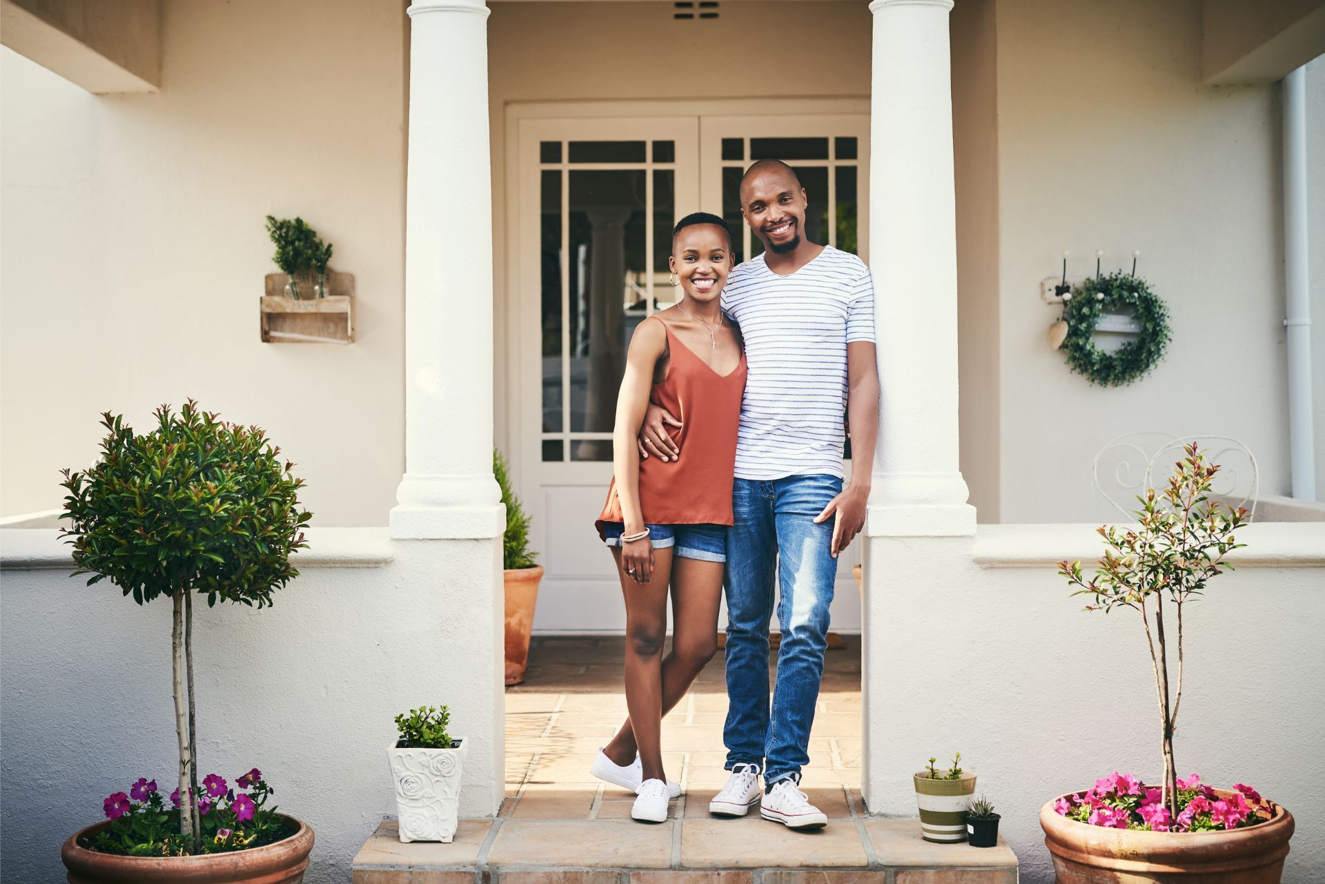 Couple standing on a porch, smiling in front of a white house with columns and potted plants.