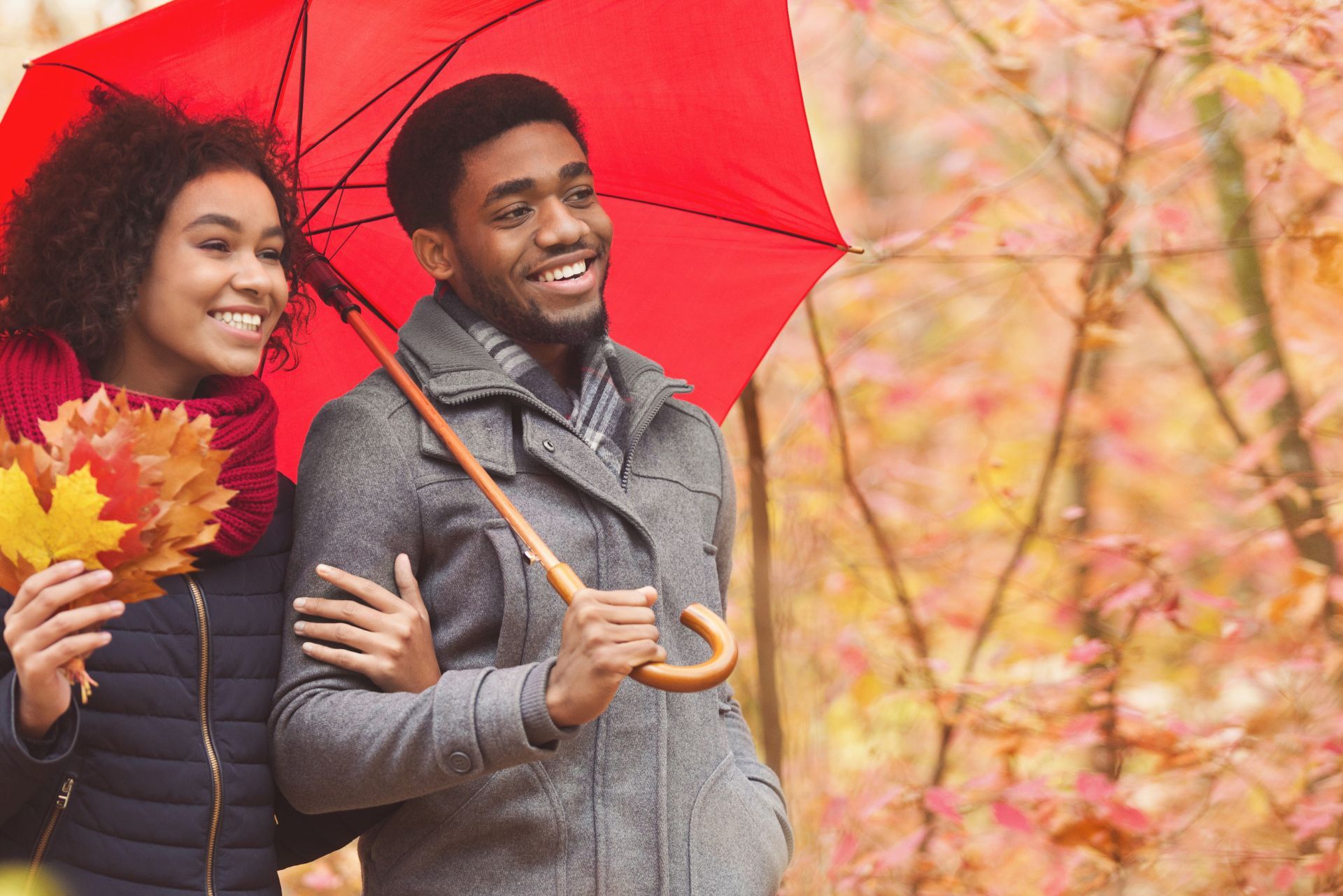 Two people smiling with a red umbrella in an autumn park, holding fall leaves.