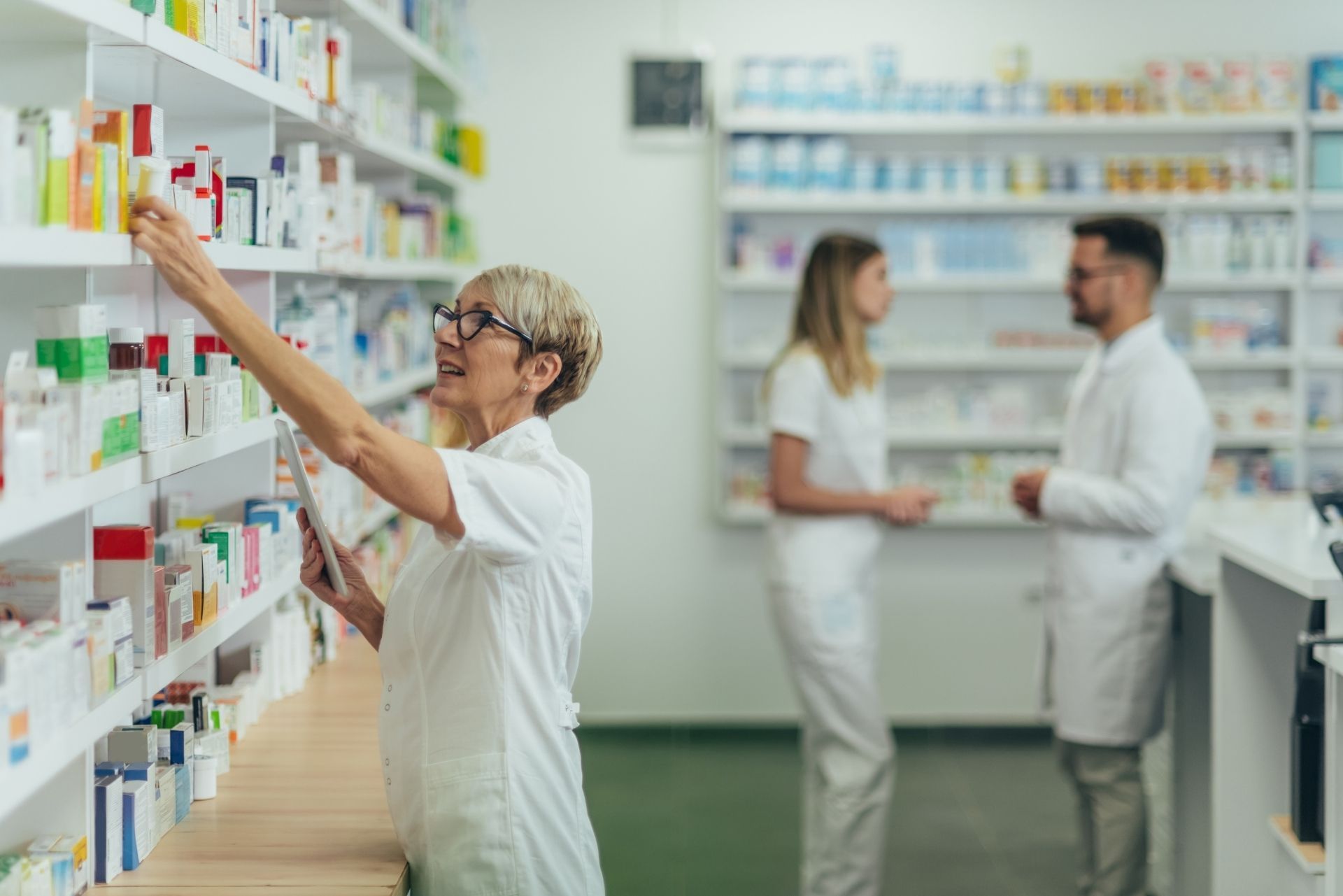 A pharmacist in a white coat reaches for medication on a shelf in a pharmacy, with colleagues standing in the background.
