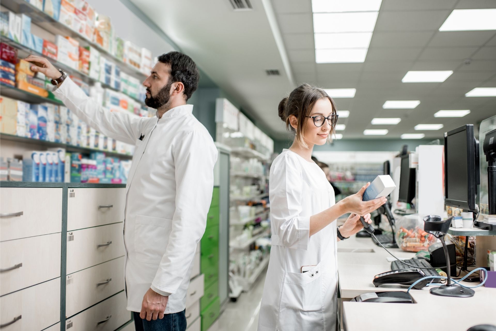 Two pharmacists in white lab coats work in a pharmacy, one retrieving items from a shelf and one reviewing a product.