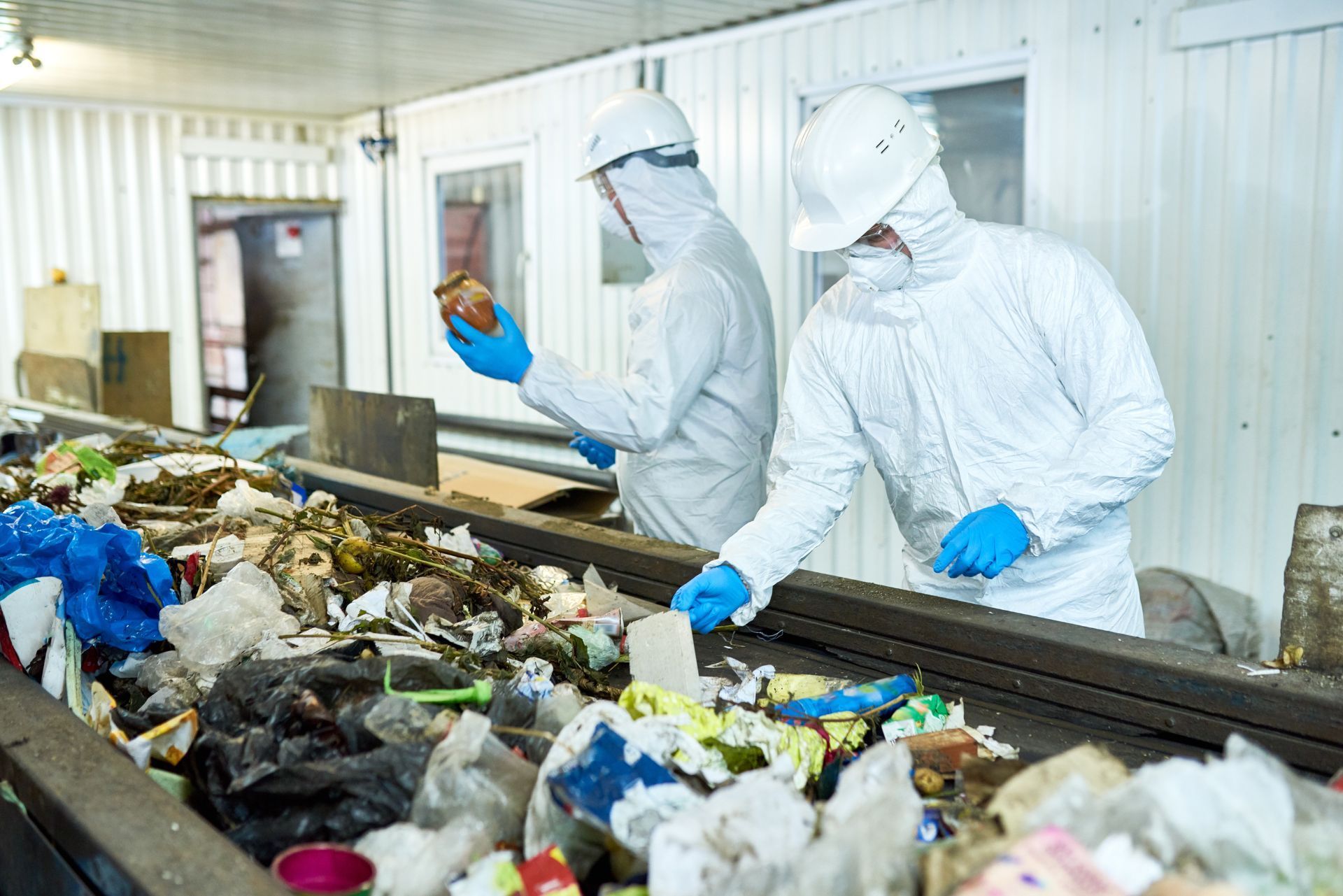 Two people in white protective suits and masks sort through garbage on a conveyor belt at a waste processing facility.