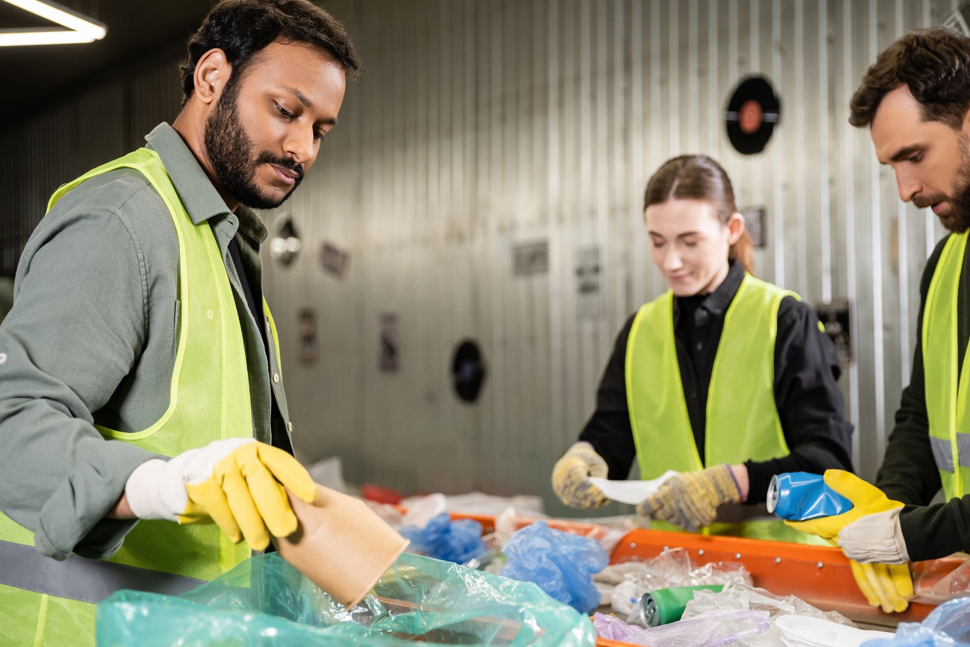 Workers in high-visibility vests sort plastic waste on a conveyor belt in a recycling facility.