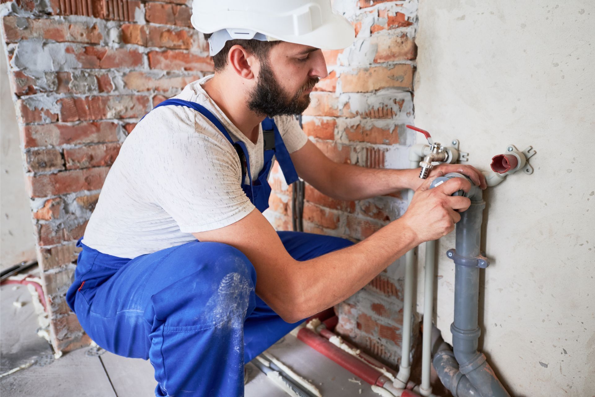 A worker in a white hard hat and blue overalls kneels while installing pipes against an exposed brick wall.